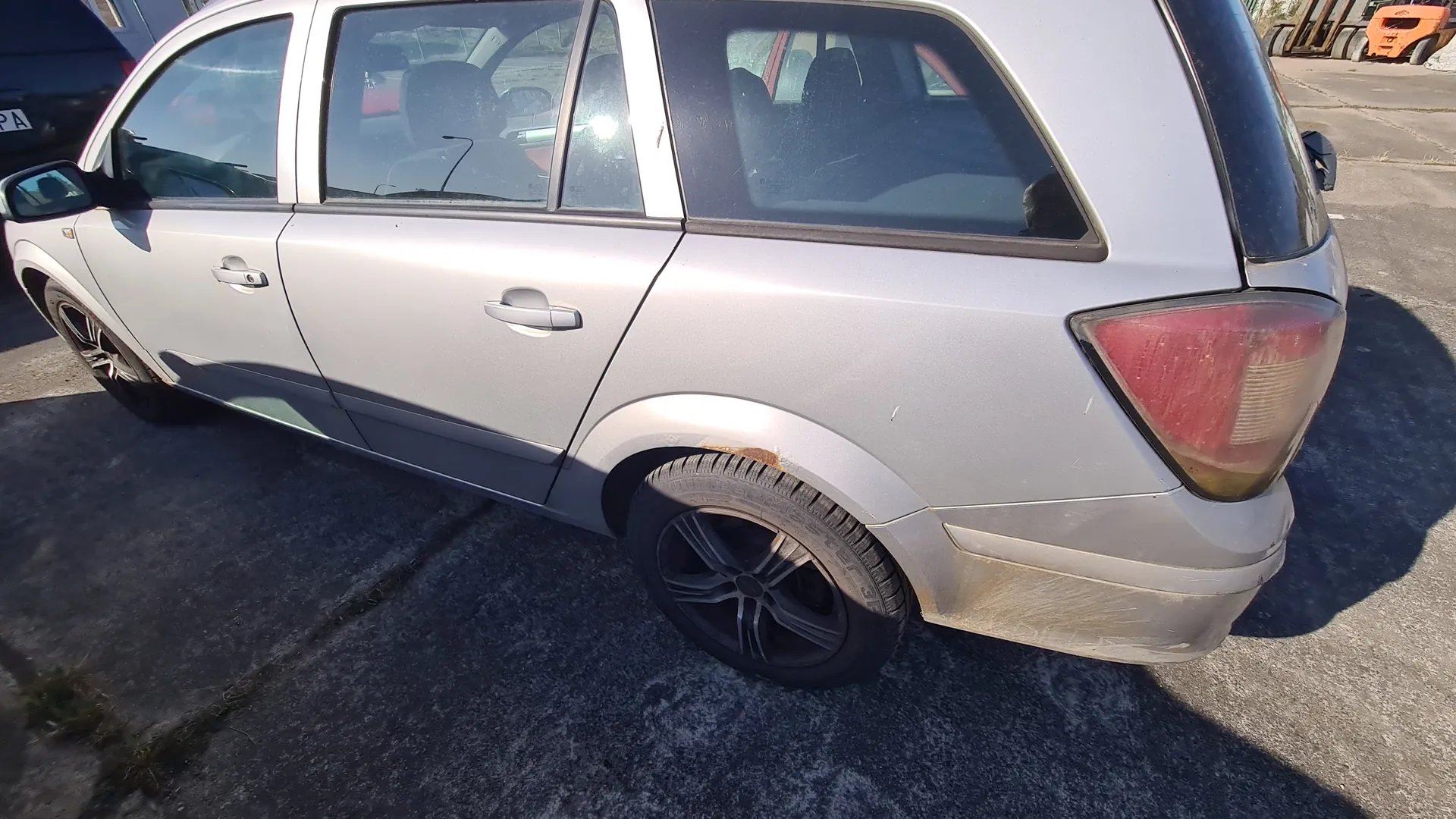 Srebrny samochód typu kombi z czterema drzwiami, widok z boku i tyłu. Widoczne są czarne felgi, czerwone światło tylnego reflektora oraz rdza na nadkolu koła tylnego. Samochód stoi na asfaltowej powierzchni. W tle częściowo widoczny inny pojazd z tablicą rejestracyjną zawierającą fragment "PA" oraz pomarańczowe maszyny budowlane.