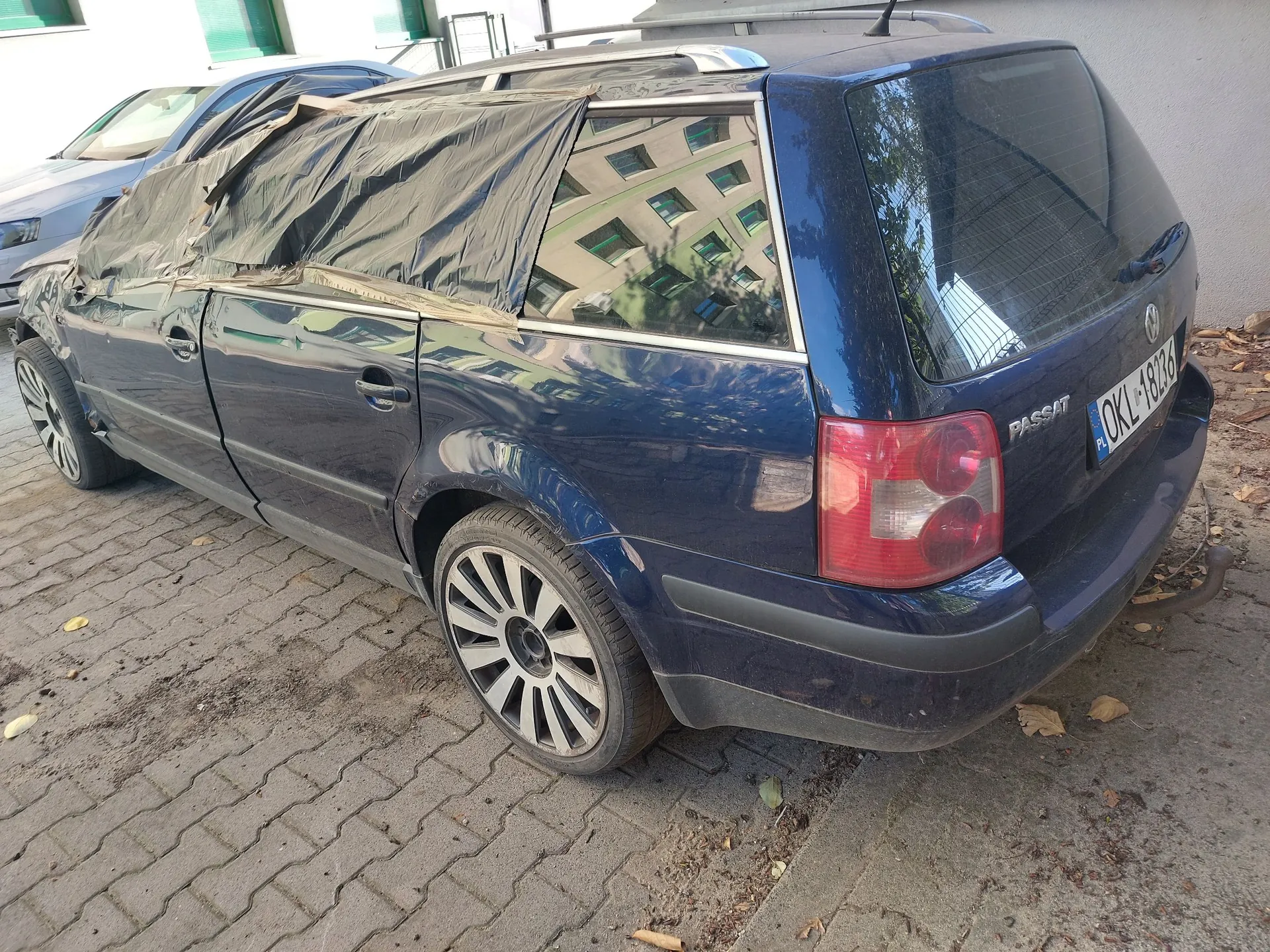 Niebieski samochód marki Volkswagen Passat w nadwoziu kombi, zaparkowany na kostce brukowej. Dach oraz tylna część samochodu są przykryte czarną, plastikową folią zabezpieczającą, która jest przymocowana taśmą. Pojazd posiada polską tablicę rejestracyjną o numerze OKL 18236. W tle widoczny jest fragment innego, jasnego samochodu oraz ściana budynku.