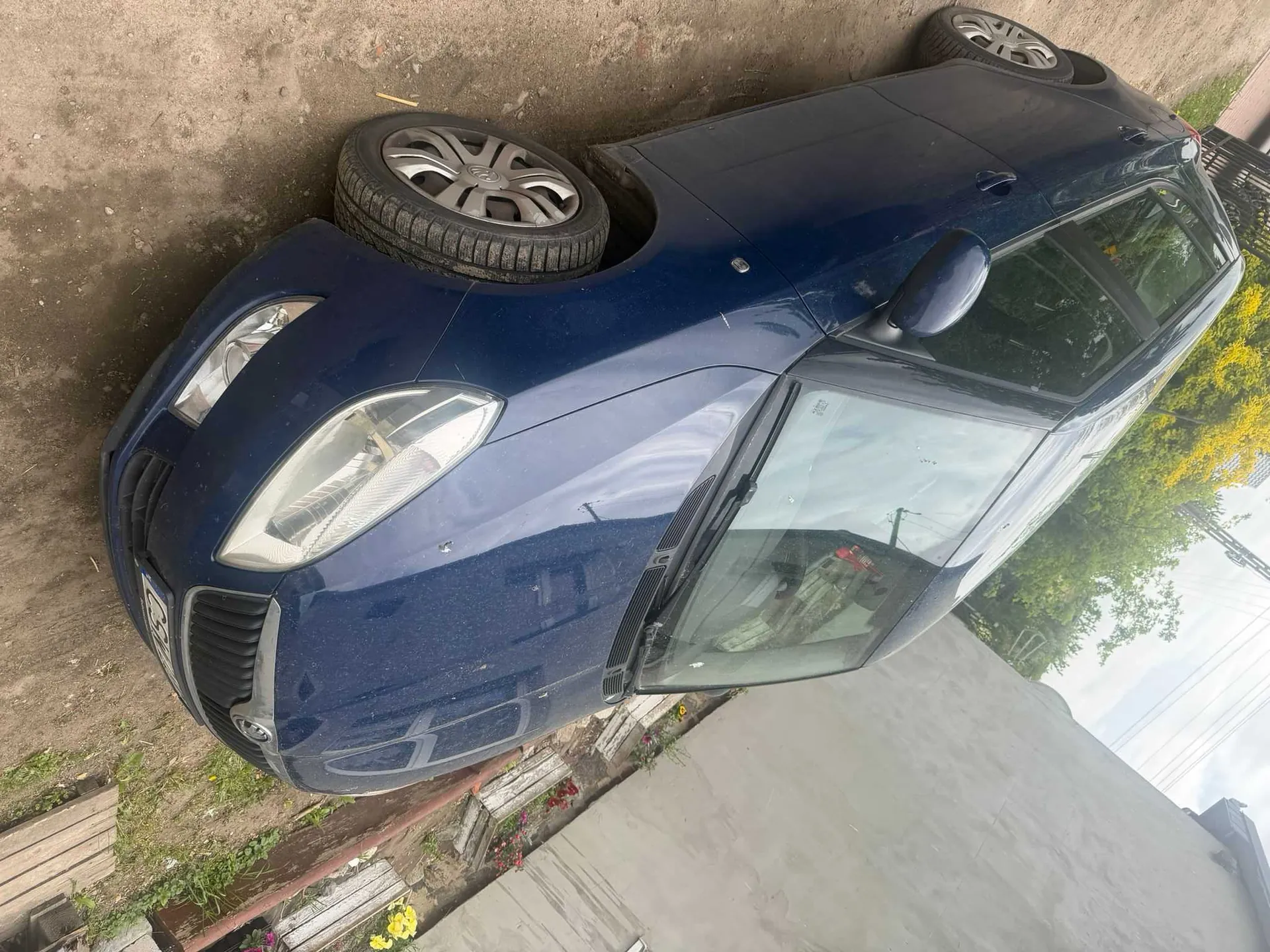 Niebieski samochód marki Skoda zaparkowany na brudnej, ziemistej powierzchni. Po prawej stronie widoczna szara ściana budynku z kwiatami w doniczkach. W tle drzewa z żółtymi kwiatami i niebo z chmurami. Samochód skierowany jest w prawą stronę obrazu, a jego przednia szyba odbija otoczenie. Widoczne są felgi i reflektory.
