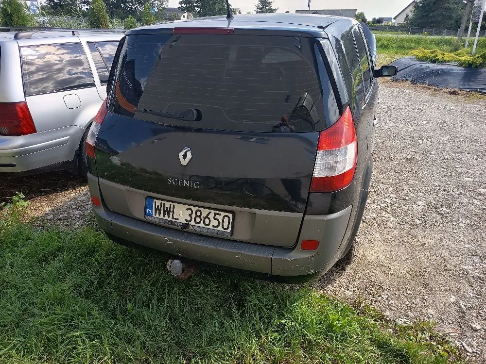 Tylny widok ciemnoszarego samochodu marki Renault Scenic zaparkowanego na żwirowym podłożu obok innego, jasnego samochodu. Na klapie bagażnika widoczne jest logo Renault, napis SCENIC oraz polska tablica rejestracyjna o numerze WWL 38650. W tle widać zieloną roślinność i fragmenty budynków.