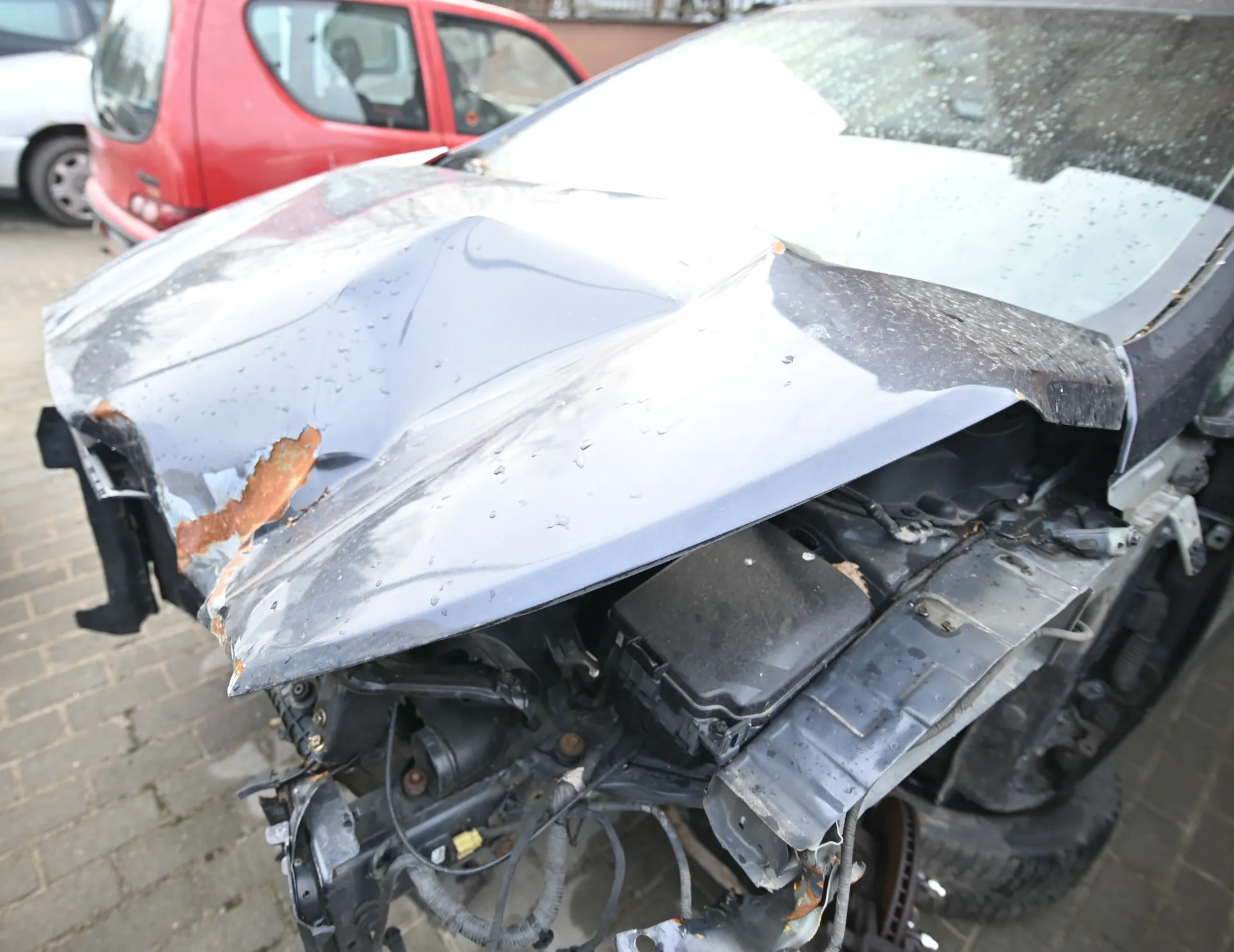 Zniszczony samochód z deformowaną maską, widoczną rdzą po lewej stronie i odsłoniętym silnikiem. Przednią szybę pokrywają pęknięcia. W tle czerwony i biały samochód zaparkowane na brukowanej powierzchni.