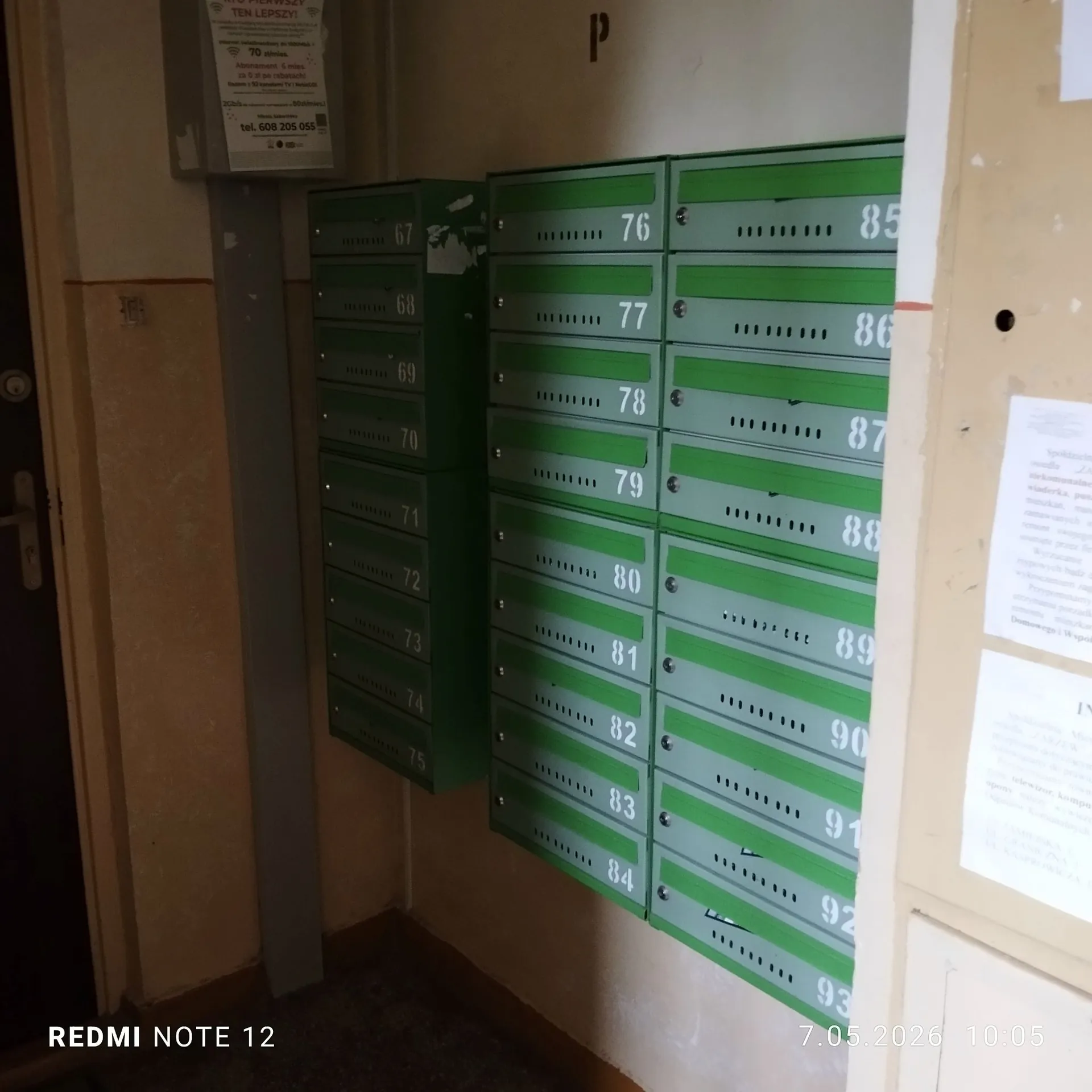 Zestaw metalowych skrzynek pocztowych zamontowanych na ścianie w korytarzu. Skrzynki są podzielone na dwie kolumny: lewa kolumna zawiera skrzynki od numeru 67 do 75, a prawa kolumna, w kolorze zielonym, zawiera skrzynki od numeru 76 do 93. Nad skrzynkami wisi biała kartka z tekstem i numerem telefonu: tel. 608 205 055. Po prawej stronie widoczna jest część białej tablicy informacyjnej z tekstem.