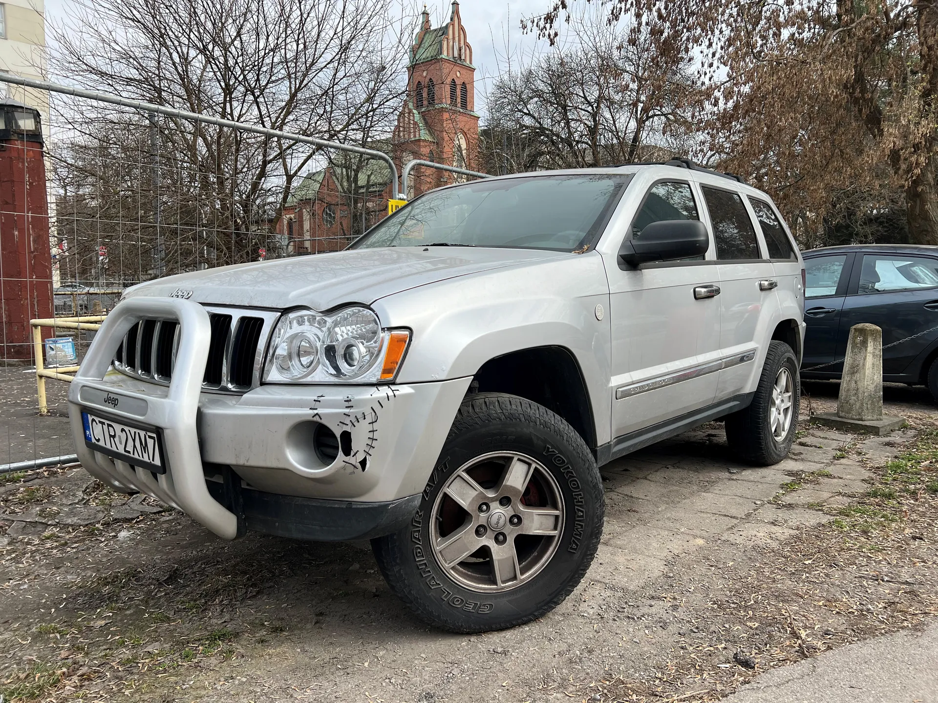 Srebrny SUV Jeep z tablicą rejestracyjną CTR 2XM7 zaparkowany na brukowanym obszarze. W tle ceglany kościół z wieżą, drzewa bez liści, metalowe ogrodzenie oraz czarny samochód osobowy. Na przednim zderzaku widoczne są ozdobne szwy. Opony marki Yokohama Geolandar A/T. Tło obejmuje również betonowy słup i suche liście na ziemi.