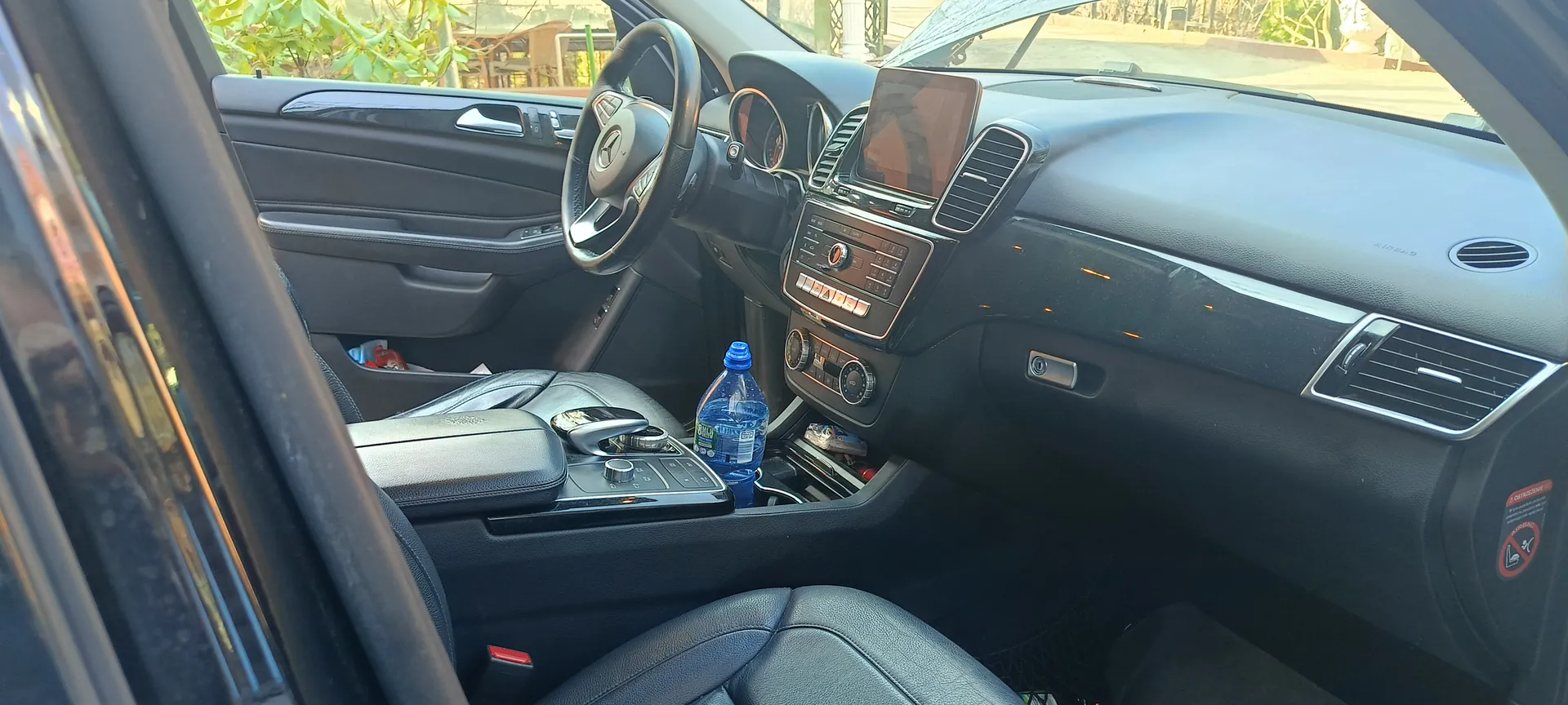 Wnętrze samochodu z widocznym kierownicą z logo, deska rozdzielcza z ekranem, klimatyzacją i przyciskami, centralna konsoła z butelką wody, czarnymi skórzonymi siedzeniami oraz otwartą drzwiami. Przez okno widać zielone rośliny i budynki na zewnątrz.