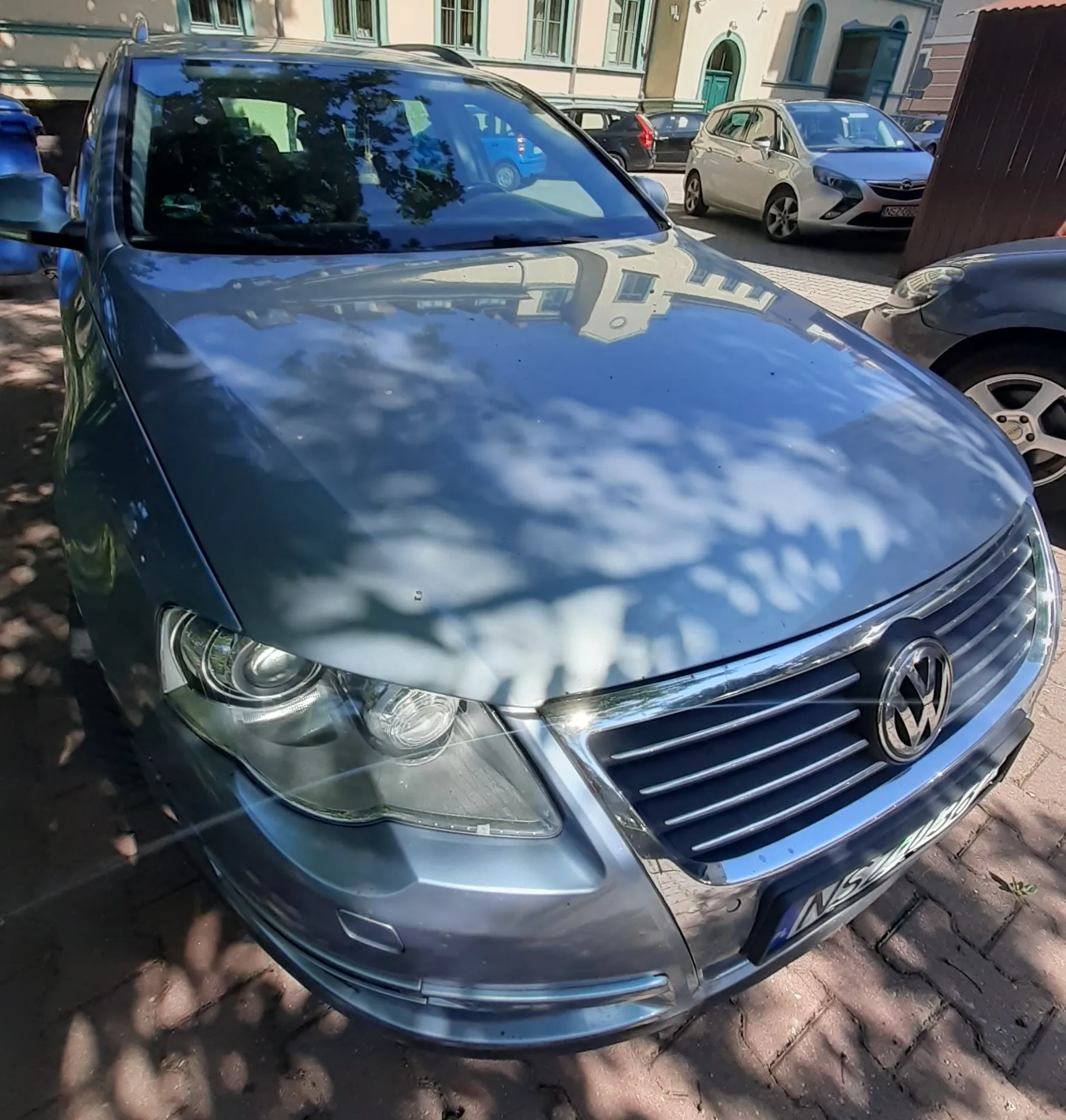 Srebrny samochód marki Volkswagen zaparkowany na kostce brukowej. Zdjęcie przedstawia przód pojazdu z widocznym logo VW na grillu oraz reflektorem. Na masce samochodu odbijają się liście drzew i okoliczne budynki. W tle widoczne są inne zaparkowane samochody oraz fasada kamienicy.