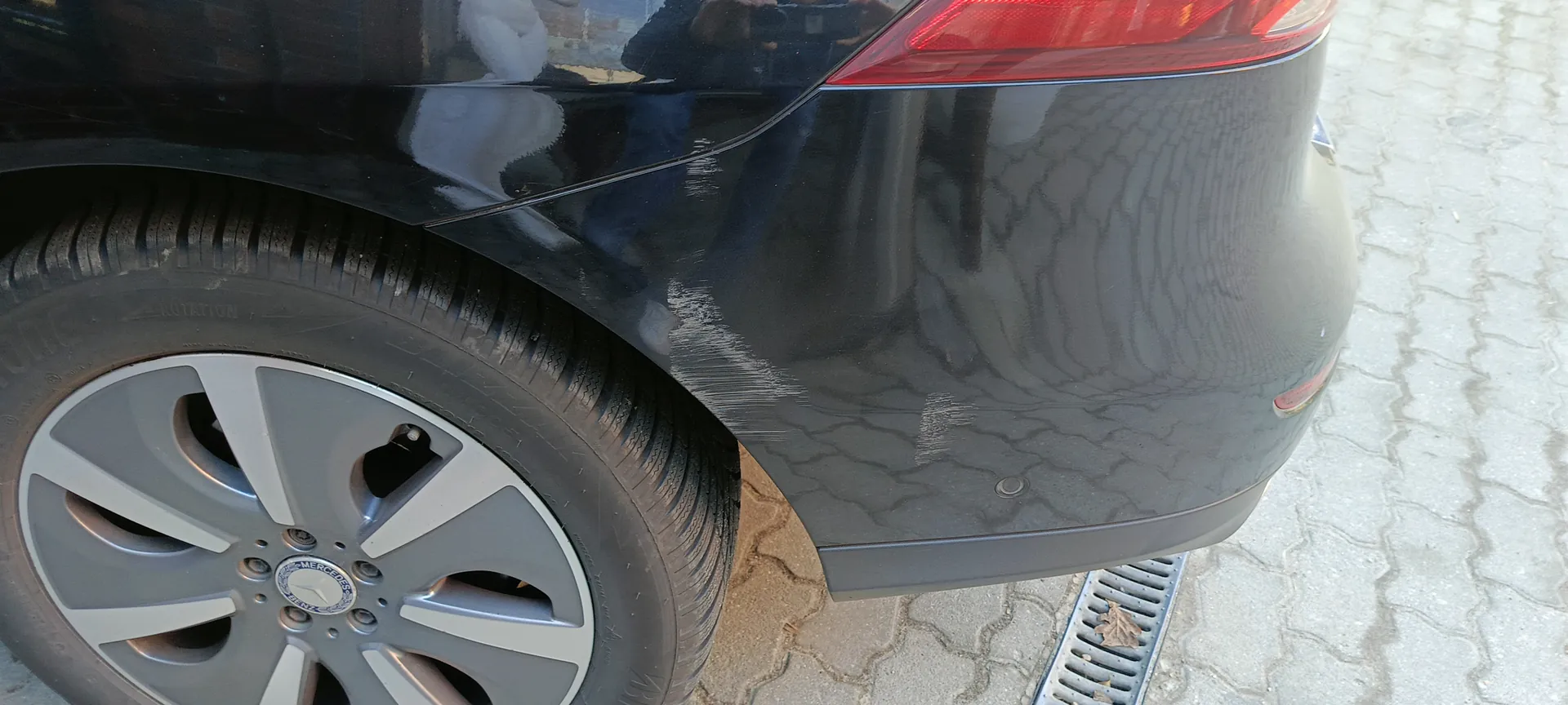 Czarna tylna część samochodu z widocznym kołem z felgą Mercedes-Benz, czerwonym światłem tylnym i odbiciami na karoserii. Samochód zaparkowany na brukowanej nawierzchni z kanałem odpływowym.