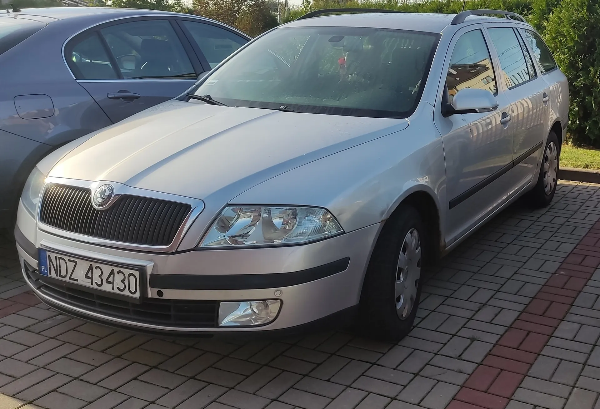 Srebrny samochód marki Skoda w nadwoziu kombi zaparkowany na kostce brukowej. Widoczna jest przednia część pojazdu z tablicą rejestracyjną o numerze NDZ 43430. Obok stoi inny, ciemnoszary samochód.