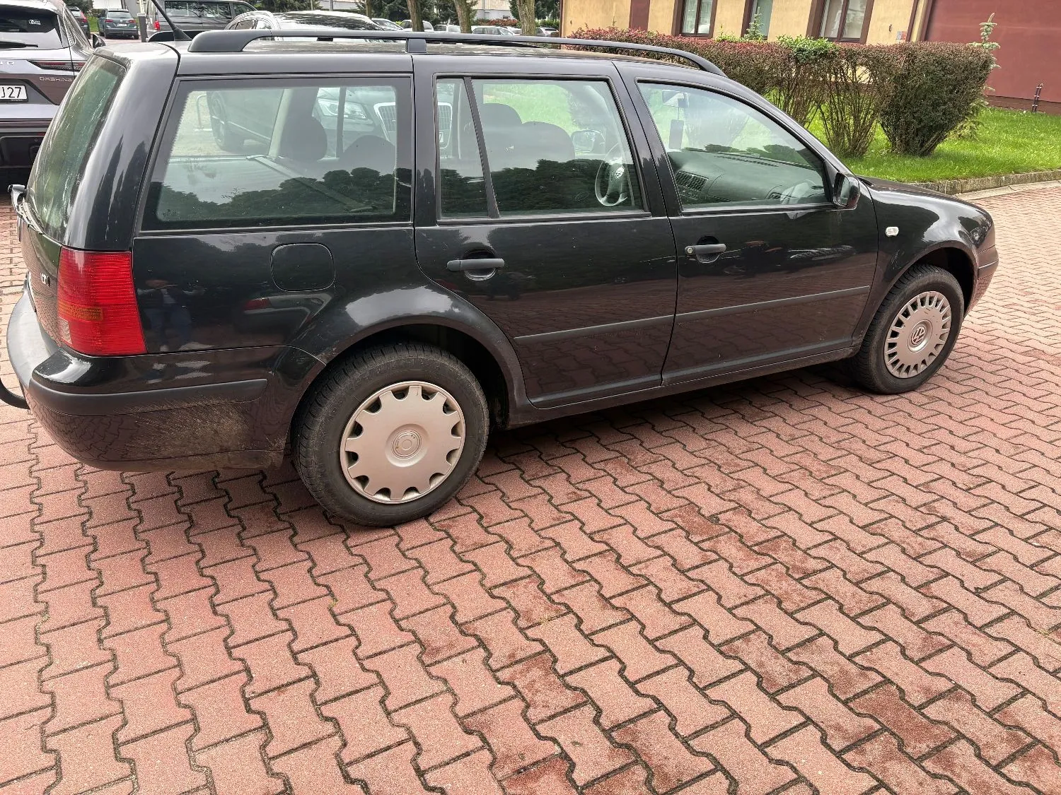 Czarny samochód typu kombi zaparkowany na podjeździe z czerwonej kostki brukowej. Pojazd jest widoczny z profilu od strony pasażera. W tle widać zielone krzewy oraz fragment budynku.