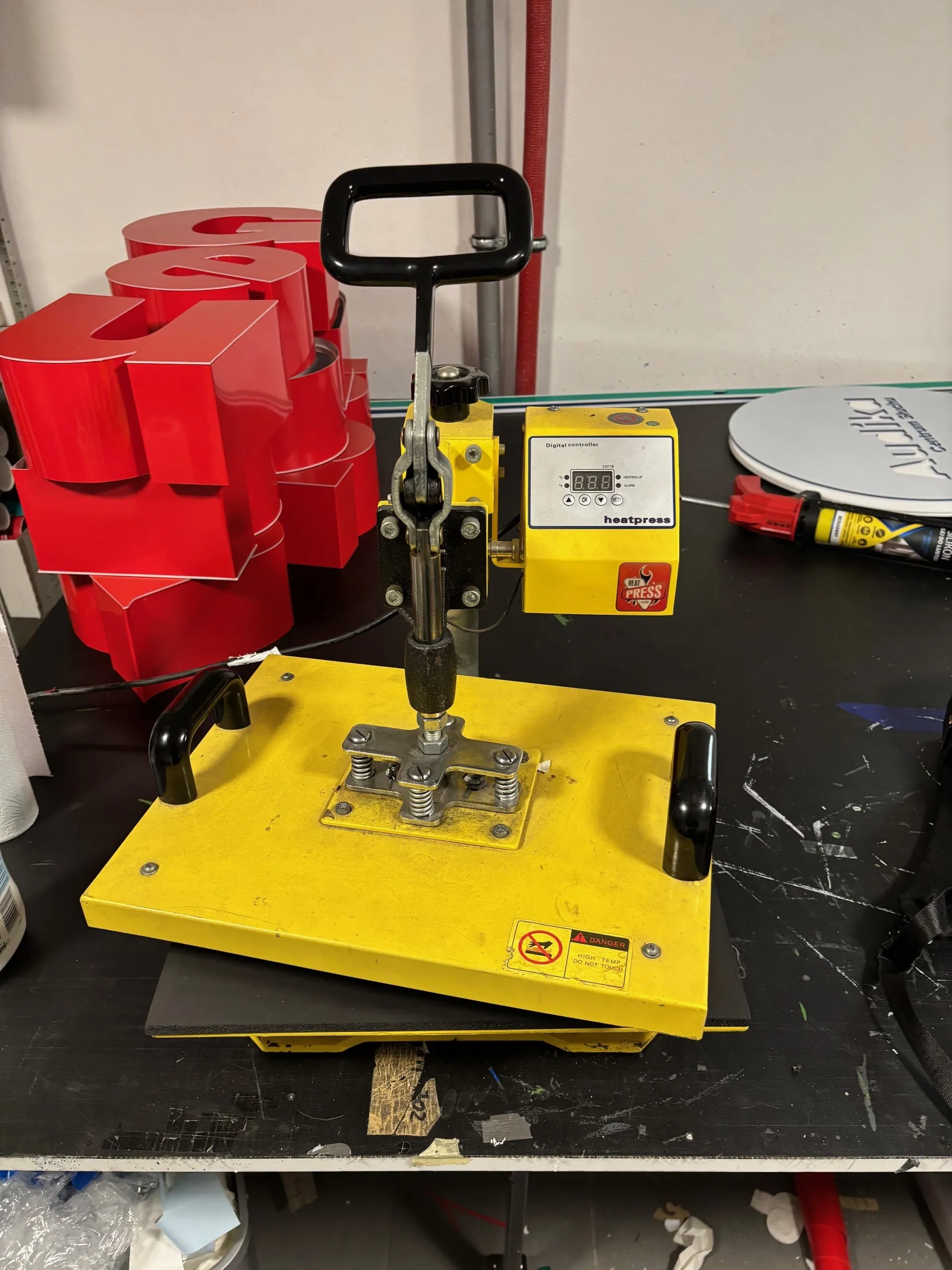 Żółty pras termiczny z czarną rączką i cyfrowym kontrolerem oznaczonym napisem 'heatpress'. Na kontrolerze wyświetlacz z cyframi '8.8.8.8' oraz przyciskami. Na podstawie prasu naklejka ostrzegawcza z symbolem zakazu i napisem 'DANGER'. W tle czerwone trójwymiarowe litery, czarna powierzchnia robocza z zadrapaniami oraz okrągły przedmiot z częściowo widocznym napisem. Obok prasu narzędzie z żółto-czerwoną obudową.