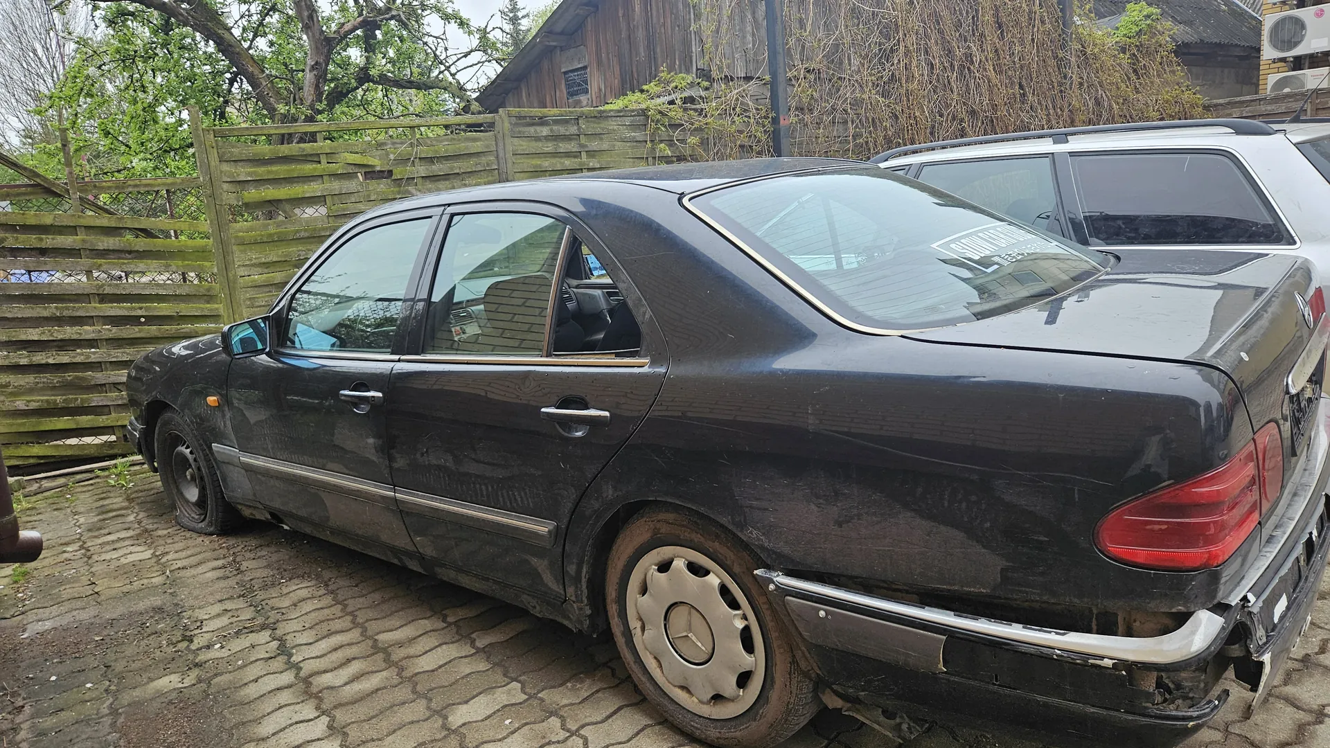 Czarny samochód osobowy typu sedan zaparkowany na brukowanej powierzchni. Pojazd ma uszkodzony tylny zderzak, brudną karoserię i koła z metalowymi felgami. W tle widoczny jest drewniany płot, drzewa, stary budynek z drewna oraz biały samochód. Na tylnej szybie czarnego samochodu znajduje się częściowo widoczny napis w języku rosyjskim. Tło obejmuje również zielone rośliny i chmurne niebo.