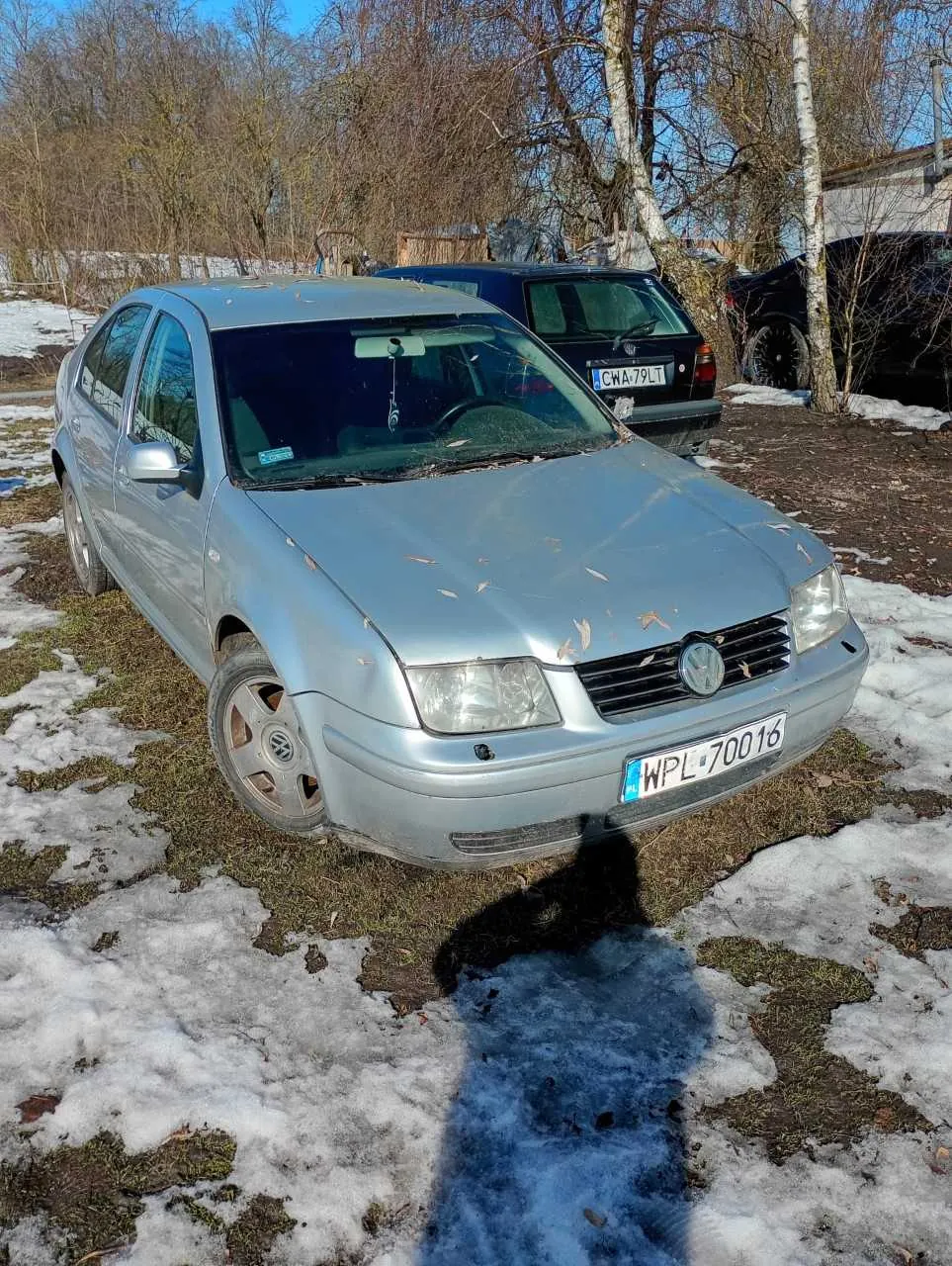 Srebrny samochód marki Volkswagen z tablicą rejestracyjną WPL 70016 na pierwszym planie. Za nim czarny samochód z tablicą CWA 79LT. Tło: zima, resztki śniegu na trawie, drzewa bez liści, niebo. Cień osoby fotografującej widoczny na śniegu.