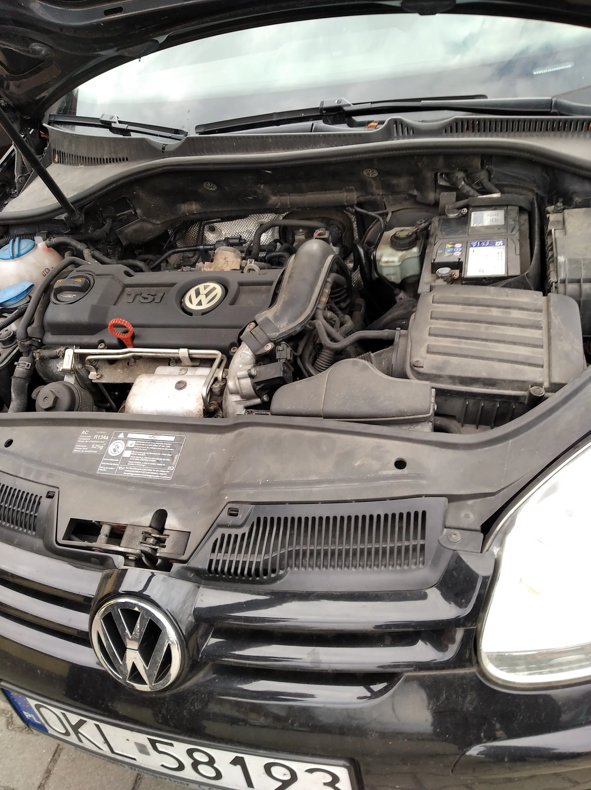 Czarny samochód marki Volkswagen z otwartą maską, ukazujący silnik z widocznym logo VW i napisem TSI. W silniku widoczny czerwony uchwyt miernika poziomu oleju. Na przedniej części pojazdu znajduje się tablica rejestracyjna z numerem OKL 58193 oraz emblemat VW na atrapie chłodnicy. W tle częściowo widoczne przednie szyby i elementy silnika, w tym zbiornik płynu chłodniczego z niebieskimi nakrętkami. Na osłonie silnika umieszczona jest czarna etykieta z białym tekstem.