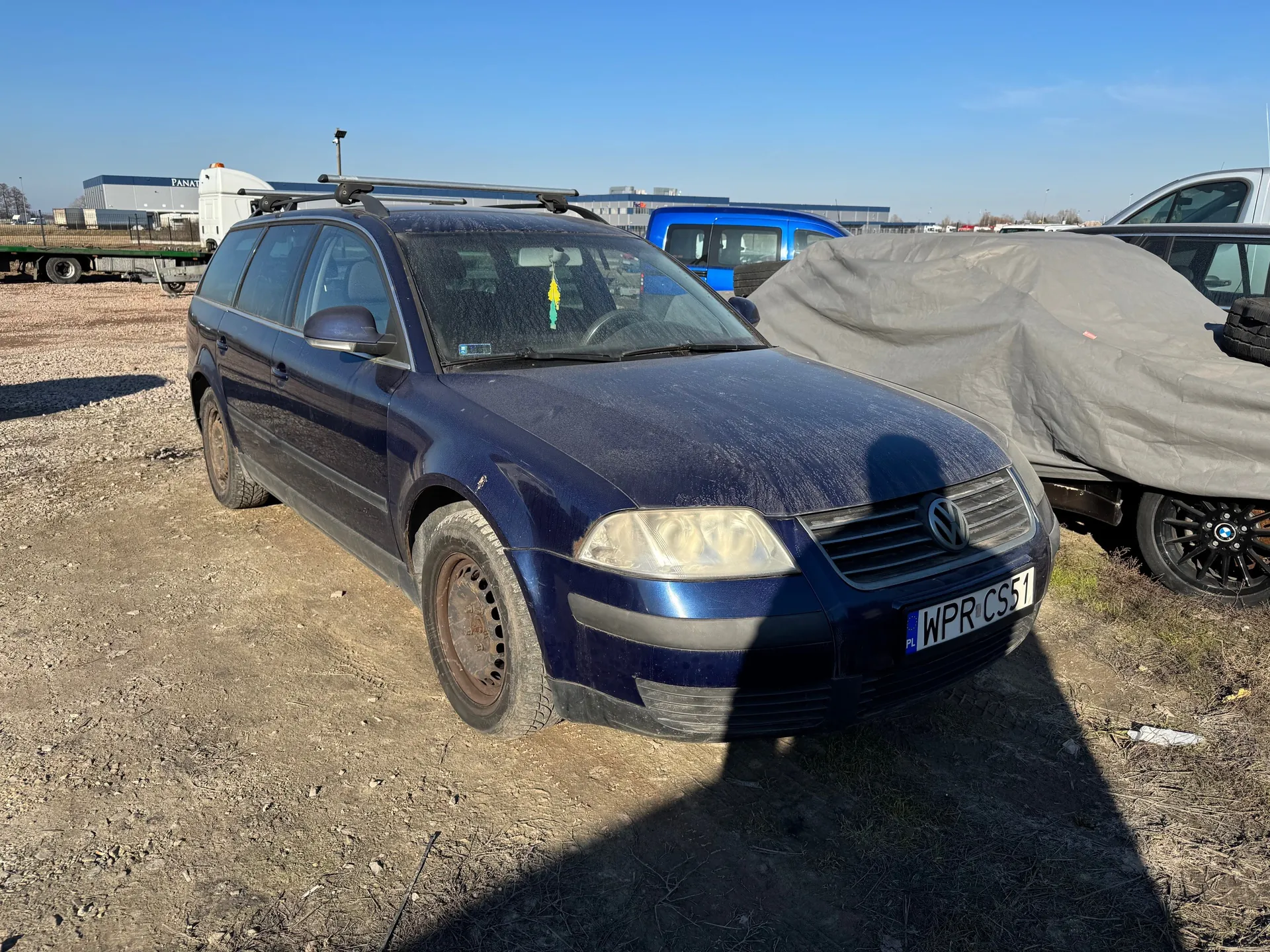 Ciemnoniebieski samochód osobowy marki Volkswagen z dachowym bagażnikiem, stojący na żwirowym placu. Widoczna tablica rejestracyjna WPR CS51 z oznaczeniem PL. W tle niebieski van, zakryty szarą pokrywą pojazd, przemysłowe budynki oraz jasne niebo. Na przedniej szybie wiszą żółto-zielone ozdoby.