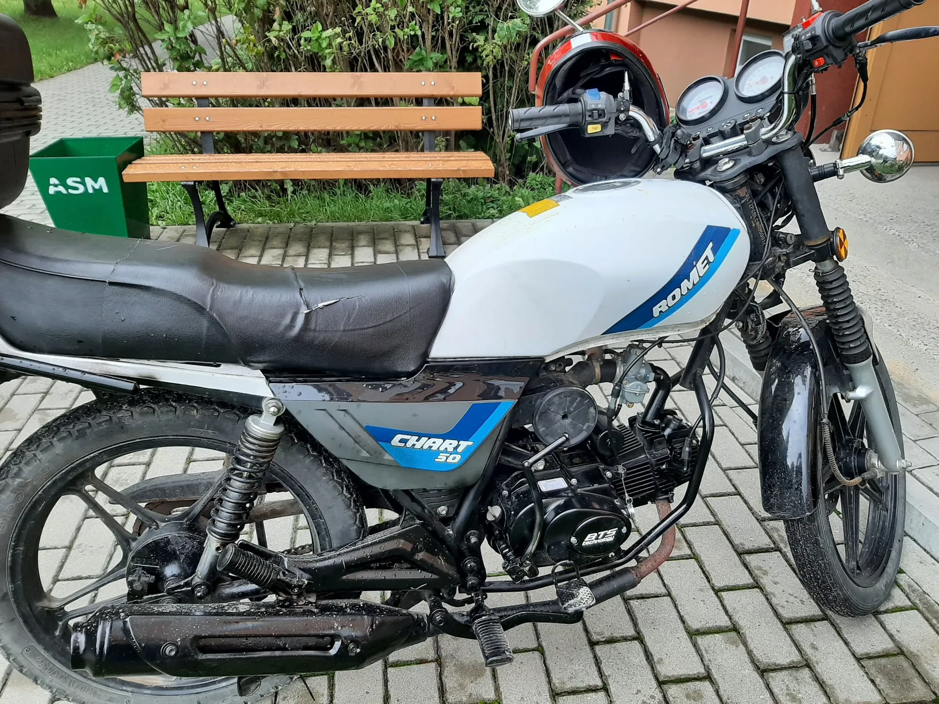 Biały motocykl marki Romet Chart 50 zaparkowany na kostce brukowej. Na baku widnieje napis 'ROMET', a na bocznej owiewce napis 'CHART 50'. W tle znajduje się drewniana ławka oraz zielony kosz na śmieci z napisem 'ASM'.