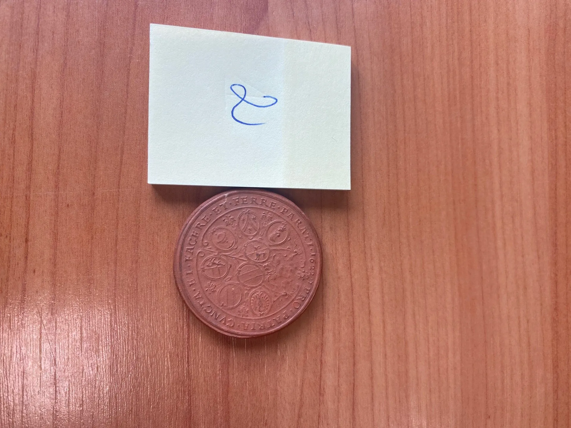 Na drewnianej powierzchni leży jasny kwadratowy arkusz papieru z niebieskim, ręcznie napisanym symbolem ampersand (&). Poniżej papieru znajduje się okrągły, brązowy medalion z inskrypcjami w języku łacińskim i dekoracyjnymi elementami. Widoczne fragmenty tekstu na medalionie to 'ET FERRE PARATI' oraz inne napisy ułożone w okrąg.