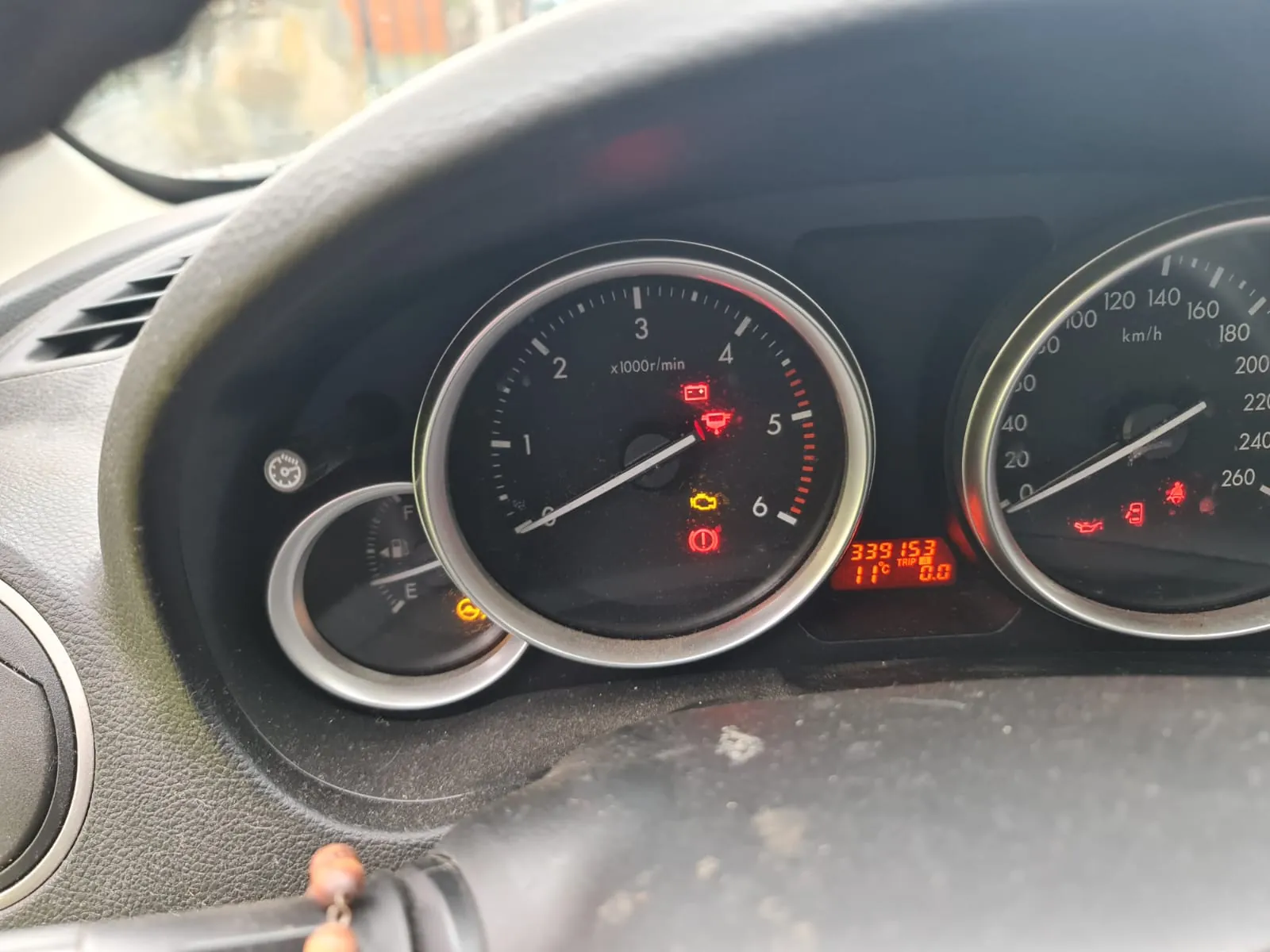 Zbliżenie na deski rozdzielcze samochodu. Widoczne są trzy wskaźniki: po lewej paliwowym (zakres E-F), w środku obrotomierz (0-6 x1000r/min) z czerwonymi i żółtymi lampkami ostrzegawczymi (m.in. akumulator, olej, silnik), po prawej prędkościomierz (0-260 km/h). Na wyświetlaczu cyfrowym czerwone cyfry: 339153, 11°C, 0.0. Fragment kierownicy i deski rozdzielczej z widocznymi elementami wnętrza pojazdu.