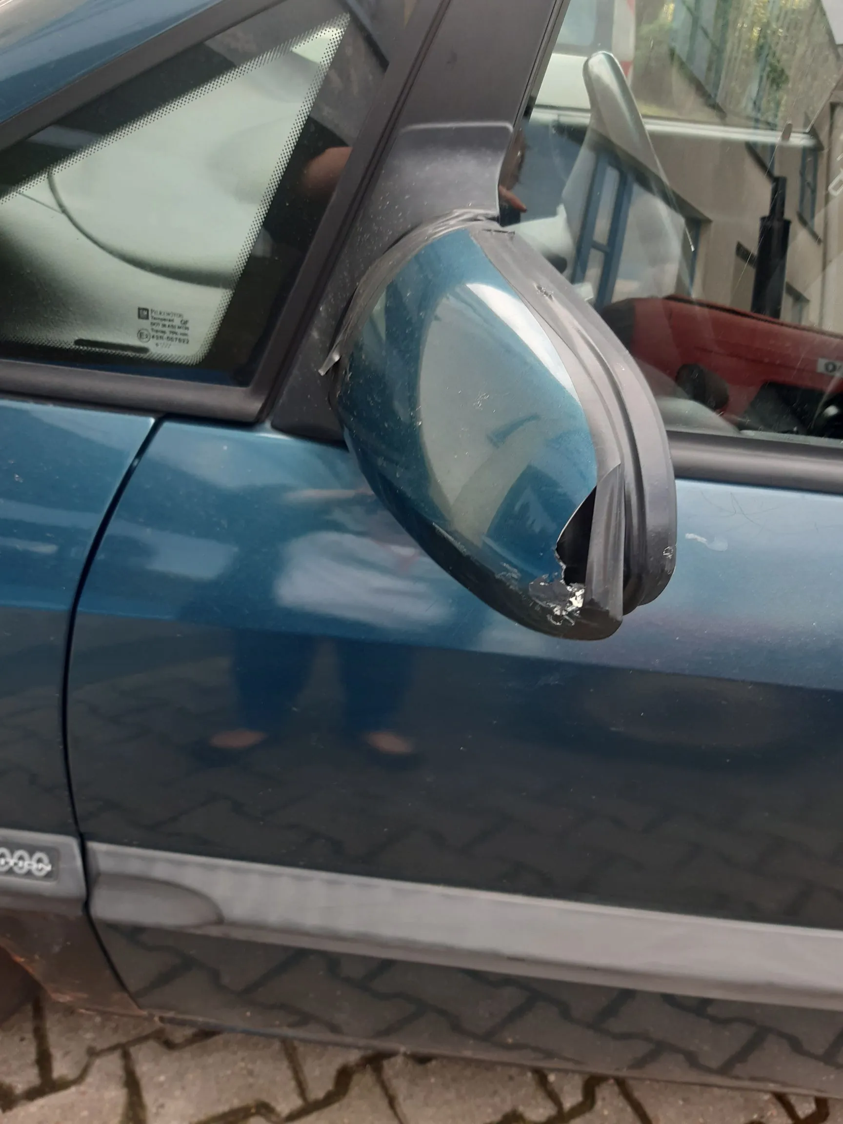 Zbliżenie na uszkodzone prawe lusterko boczne w niebieskim samochodzie. Obudowa lusterka jest pęknięta i częściowo oderwana, odsłaniając wewnętrzne elementy. W tle widoczny jest fragment drzwi samochodu z szarym paskiem ochronnym oraz fragment chodnika z kostki brukowej.