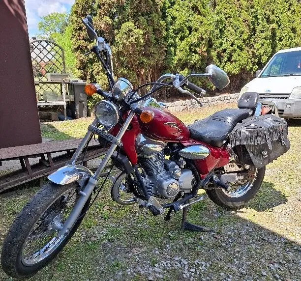 Czerwony motocykl z czarnym siedzeniem i frędzlami na bagażniku, zaparkowany na podłożu z żwiru i trawy. W tle biały samochód, zielone krzewy, drewniana konstrukcja z siatką oraz niebo z chmurami. Motocykl posiada chromowane elementy, reflektory i lusterka boczne.