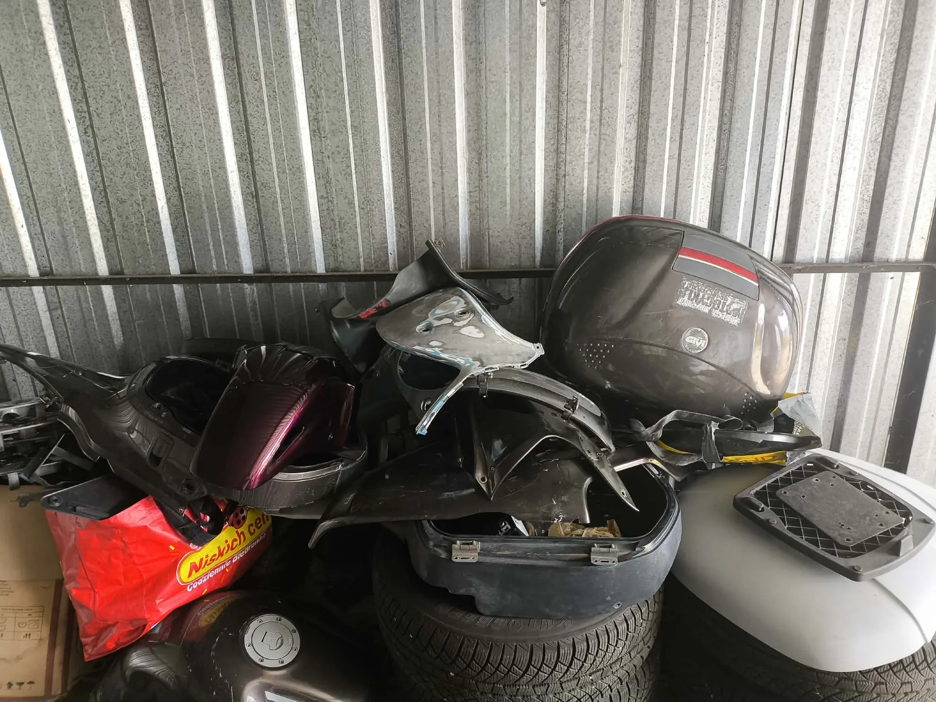 Sterta części motocyklowych i akcesoriów oparta o ścianę z blachy falistej. Wśród przedmiotów znajduje się czarny kask marki GIVI, plastikowe owiewki w kolorach czarnym i fioletowym, czarna plastikowa skrzynia, opony samochodowe oraz czerwona torba z napisem 'Nicekch cen'.