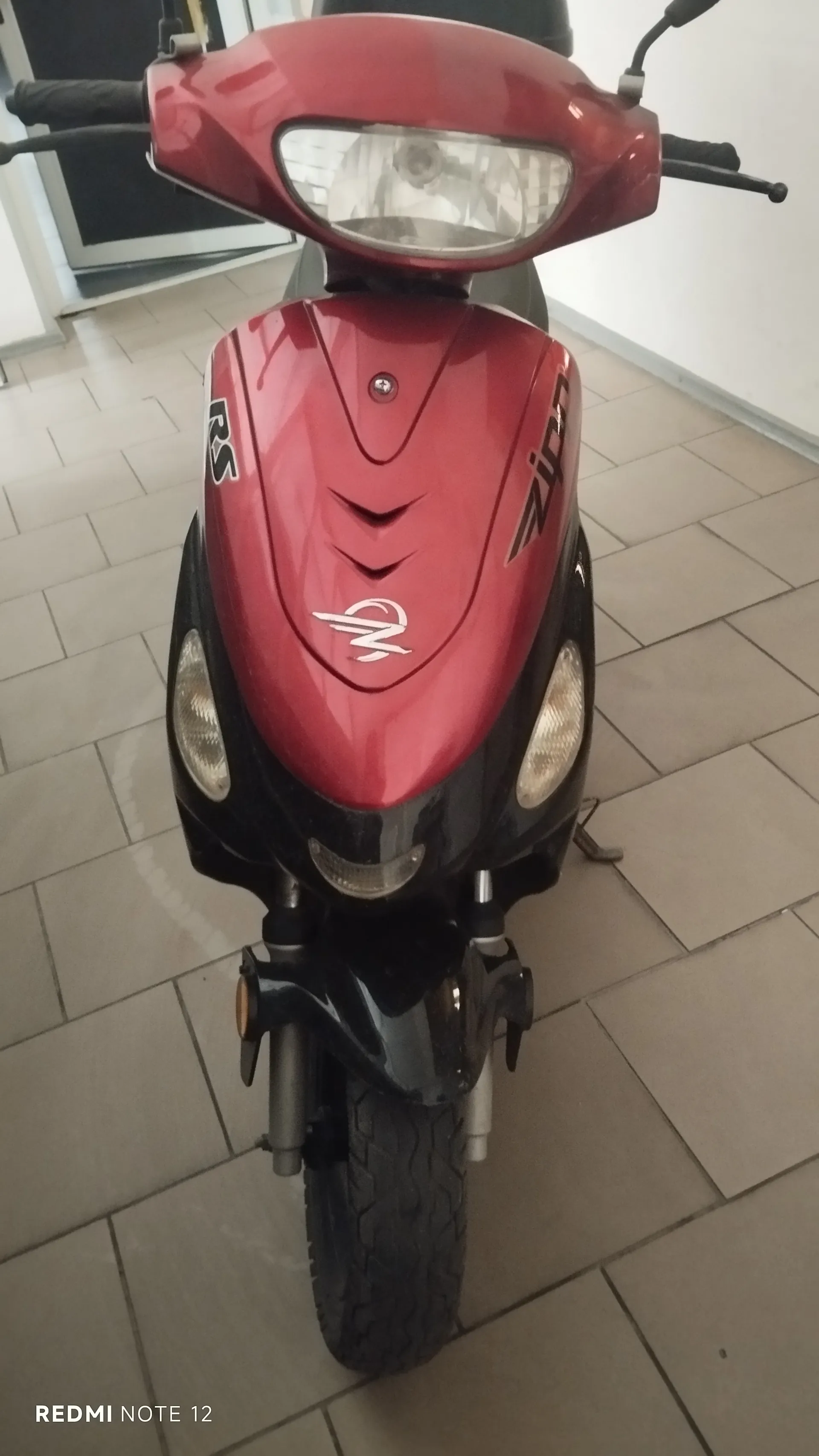 Czerwony skuter widziany od przodu z widocznym reflektorem, kierownicą i czarnymi elementami. Na czerwonej części przedniej widoczne są napisy: po lewej stronie "RS", po prawej "Zipp" oraz centralnie umieszczone logo. Tło stanowi podłoga z kafelkami i biała ściana z drzwiami. W lewym dolnym rogu obrazu znajduje się napis "REDMI NOTE 12".