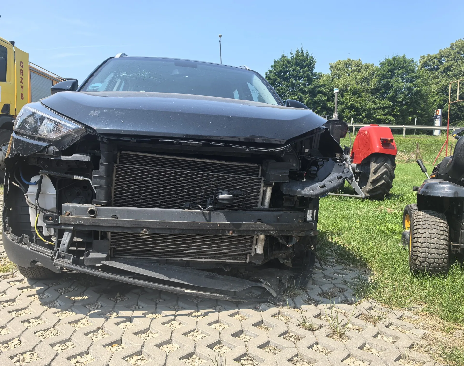 Zdjęcie przodu czarnego samochodu osobowego z poważnymi uszkodzeniami. Przedni zderzak oraz grill są całkowicie wyrwane, odsłaniając chłodnicę i elementy konstrukcyjne przodu pojazdu. Samochód stoi na kostce brukowej typu ażurowego, obok znajduje się trawnik, na którym widoczny jest fragment czerwonego traktora oraz koło innego pojazdu. W tle widać zielone drzewa i fragment żółtego pojazdu z napisem 'GRZYB'.