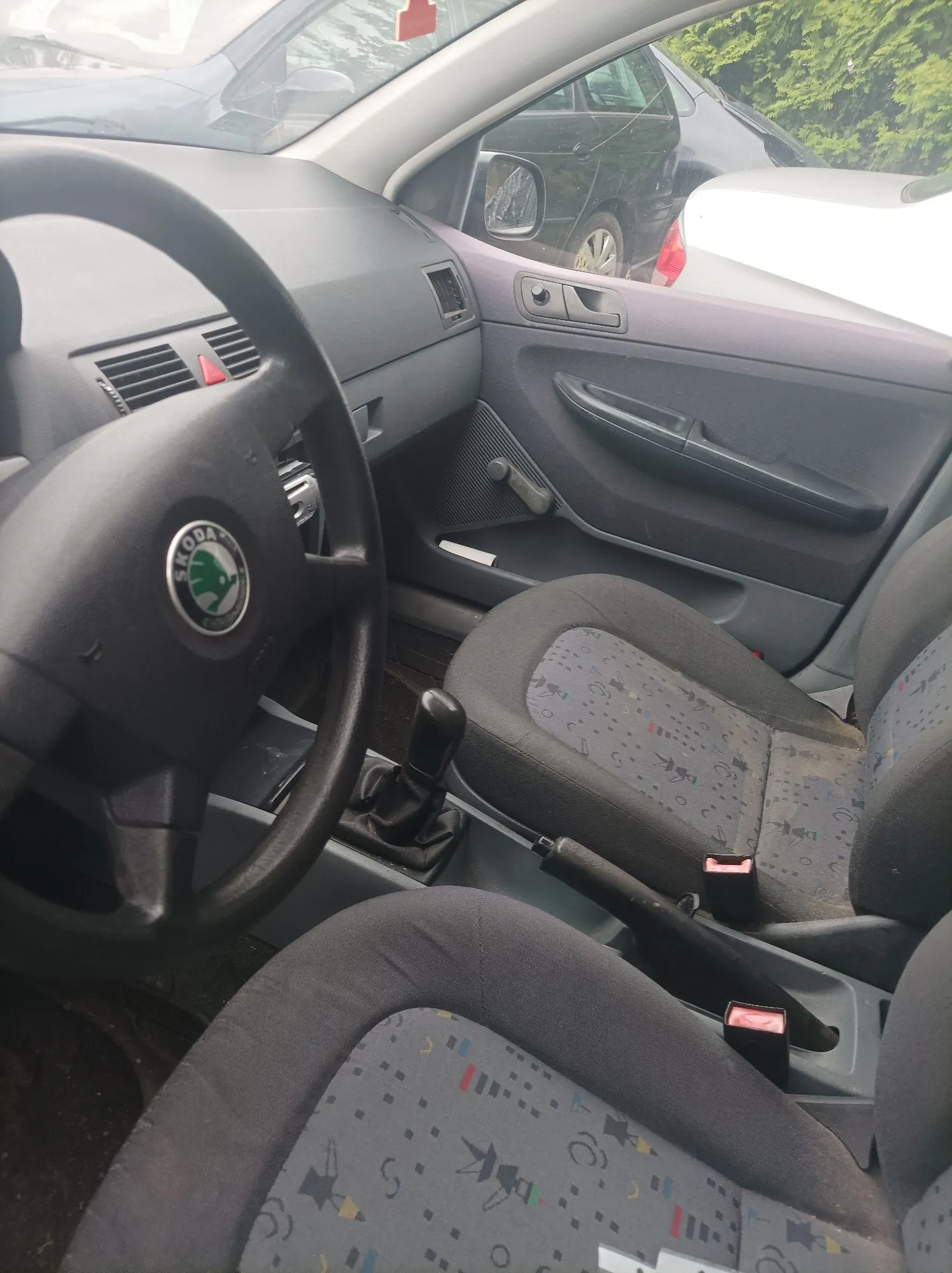 Wnętrze samochodu marki Skoda widziane z perspektywy kierowcy. Widoczna jest czarna kierownica z logo marki w centrum, lewa strona deski rozdzielczej, drzwi kierowcy oraz dwa przednie fotele obite szarym materiałem w drobny, kolorowy wzór. Pomiędzy fotelami znajduje się lewarek zmiany biegów i hamulec ręczny.