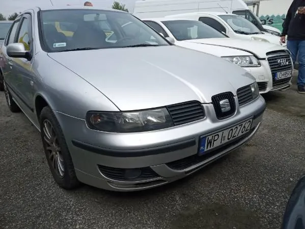 Srebrny samochód marki SEAT z tablicą rejestracyjną WP 02762 na pierwszym planie. W tle widoczne są białe samochody, w tym Audi z częściowo widoczną tablicą rejestracyjną LC. Po prawej stronie stoi osoba w ciemnej kurtce i niebieskich dżinsach. Tło stanowi parking z żwirem oraz biały van. Niebo jest pochmurne.