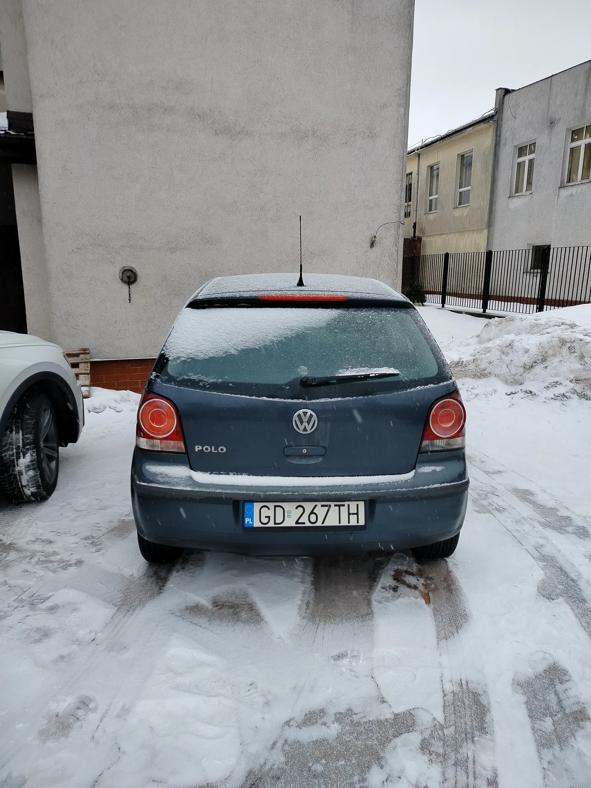 Z tyłu niebieski samochód Volkswagen Polo pokryty śniegiem, z widocznym logo VW i napisem "POLO" na bagażniku. Tablica rejestracyjna PL GD 267TH. Po lewej stronie częściowo widoczny biały samochód. Tło: szare budynki, czarna metalowa ogrodzenie i śnieżna droga z odciskami opon. Na dachu i szybie tylnej samochodu leży śnieg, a na ziemi widoczne są ślady od opon.