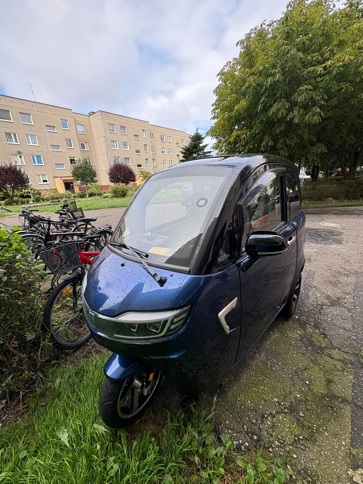 Niebieski, kompaktowy samochód elektryczny zaparkowany na poboczu drogi. Obok pojazdu stoi kilka rowerów. W tle widoczny jest wielopiętrowy blok mieszkalny z jasnej cegły oraz duże, zielone drzewa pod zachmurzonym niebem.