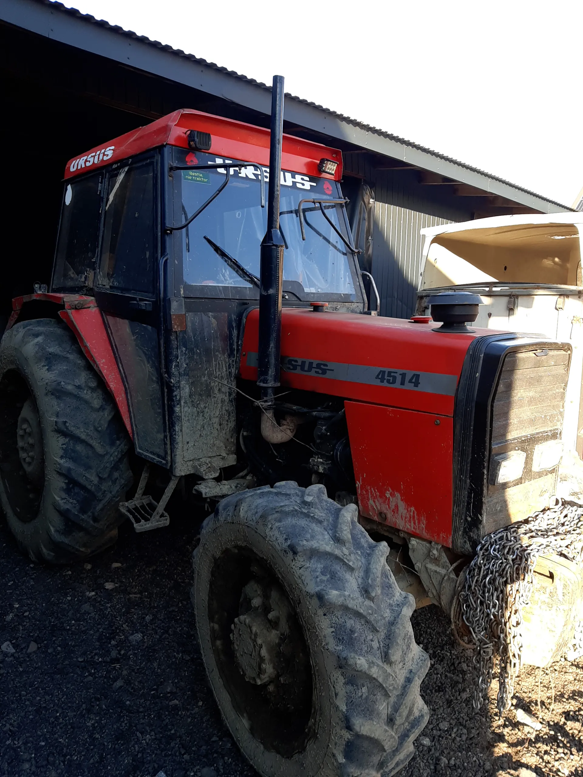 Czerwono-czarny traktor marki URSUS model 4514 z widocznym numerem 4514 na boku. Traktor ma duże, brudne opony, łańcuch umocowany na jednej z części. W tle budynek z ciemnym dachem i żwirowa powierzchnia. Tekst 'URSUS' widoczny na przodzie i boku traktora.