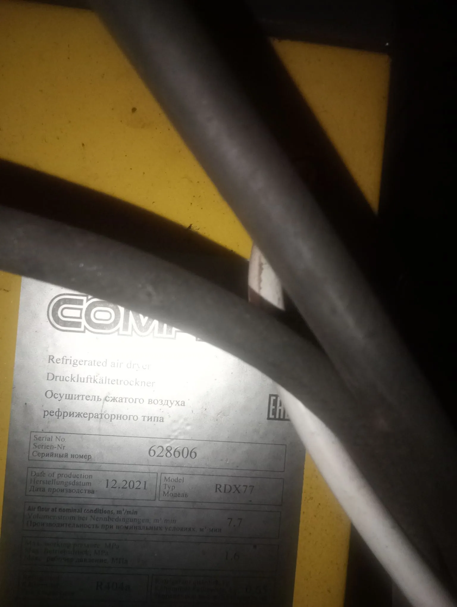 Zbliżenie na srebrną tabliczkę znamionową urządzenia przemysłowego zamontowanego na żółtej obudowie. Tabliczka zawiera tekst w języku angielskim, niemieckim i rosyjskim. Widoczne napisy to: 'Refrigerated air dryer', 'Druckluftkältetrockner', 'Осушитель сжатого воздуха рефрижераторного типа'. Podane dane techniczne to: Serial No 628606, Date of production 12.2021, Model RDX77, Air flow at nominal conditions 7.7 m³/min oraz Max working pressure 1.6 MPa. Na dole widnieje oznaczenie czynnika chłodniczego R404a. Przed tabliczką przebiegają dwa grube, czarne kable elektryczne, które częściowo zasłaniają widok.