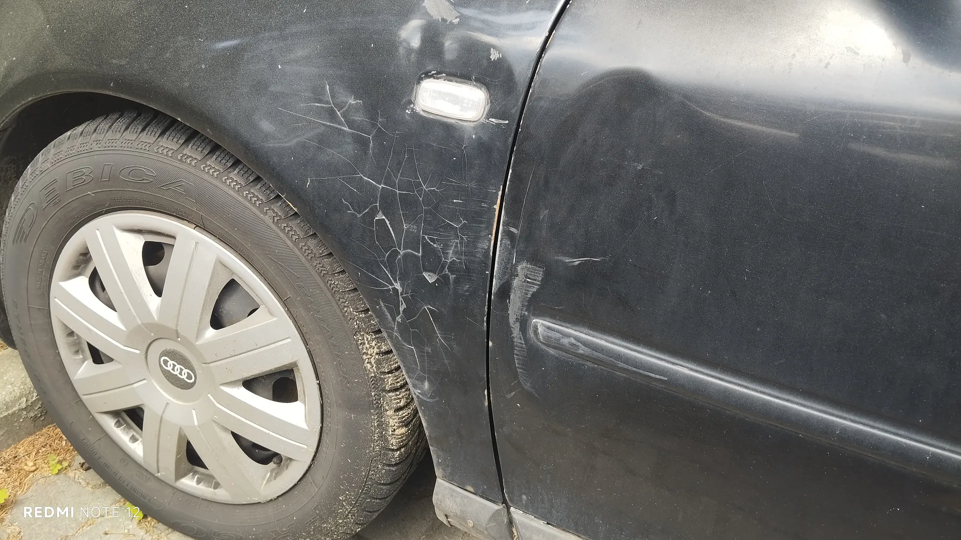 Czarny samochód marki Audi, widok na przednie koło z srebrnym felgą z logo Audi. Opona z widocznym napisem DEBICA. Na karoserii obok koła liczne zadrapania i pęknięcia lakieru. W dolnym lewym rogu obrazu widoczny napis REDMI NOTE 12. Samochód zaparkowany na betonowej powierzchni z fragmentem trawy.