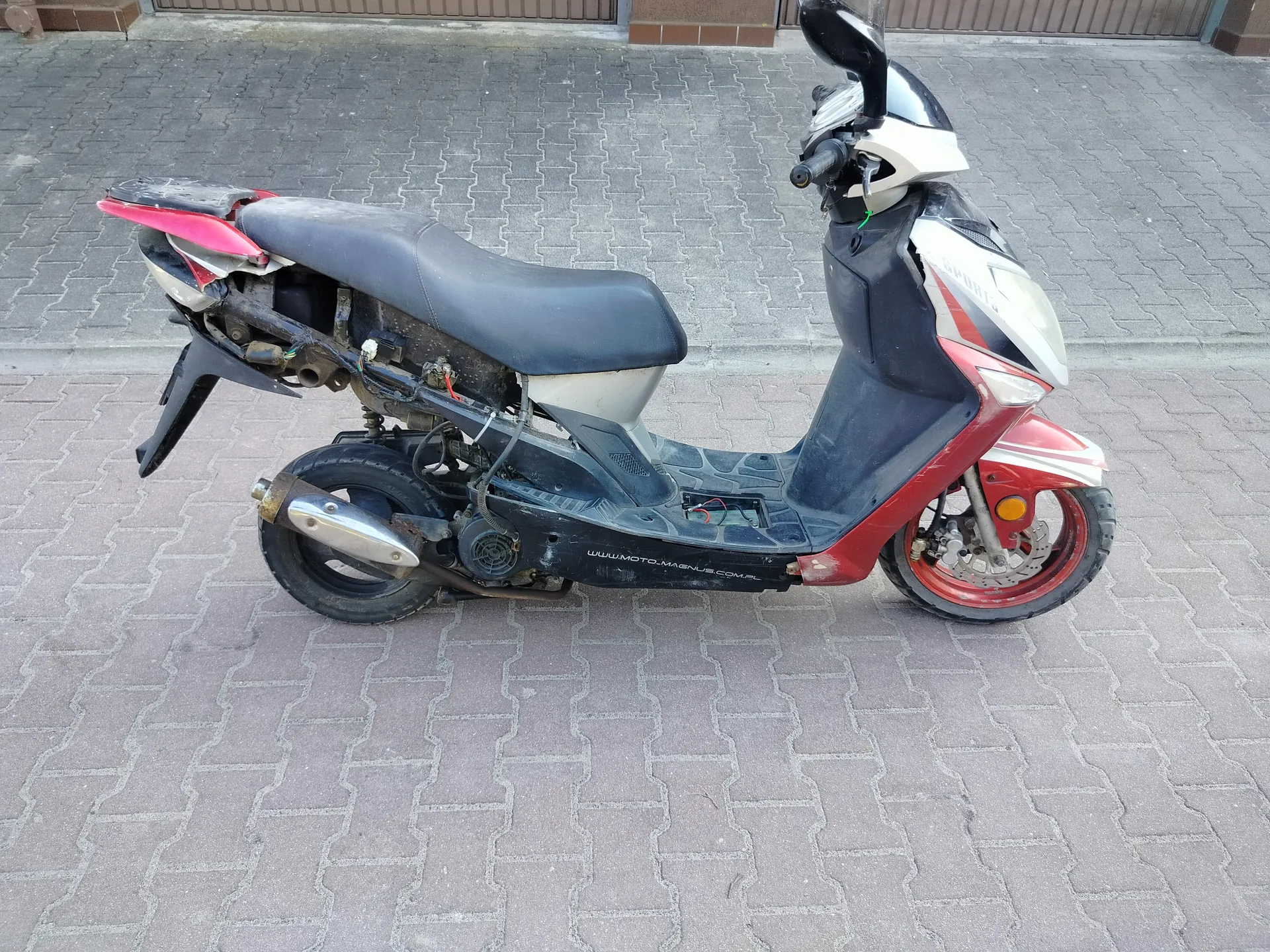Uszkodzony skuter w kolorze czerwono-czarnym z białymi akcentami, zaparkowany na brukowanej powierzchni. Widoczne uszkodzenia w tylnej części, gdzie brakuje osłony, odsłaniając elementy mechaniczne i przewody. Czarna siedzisko, czerwone felgi, reflektor przedni oraz lusterko wsteczne. Na boku skutera widoczny napis www.moto-magnus.com.pl. Tło stanowi brukowana ulica i fragment budynku z brązowym ogrodzeniem.