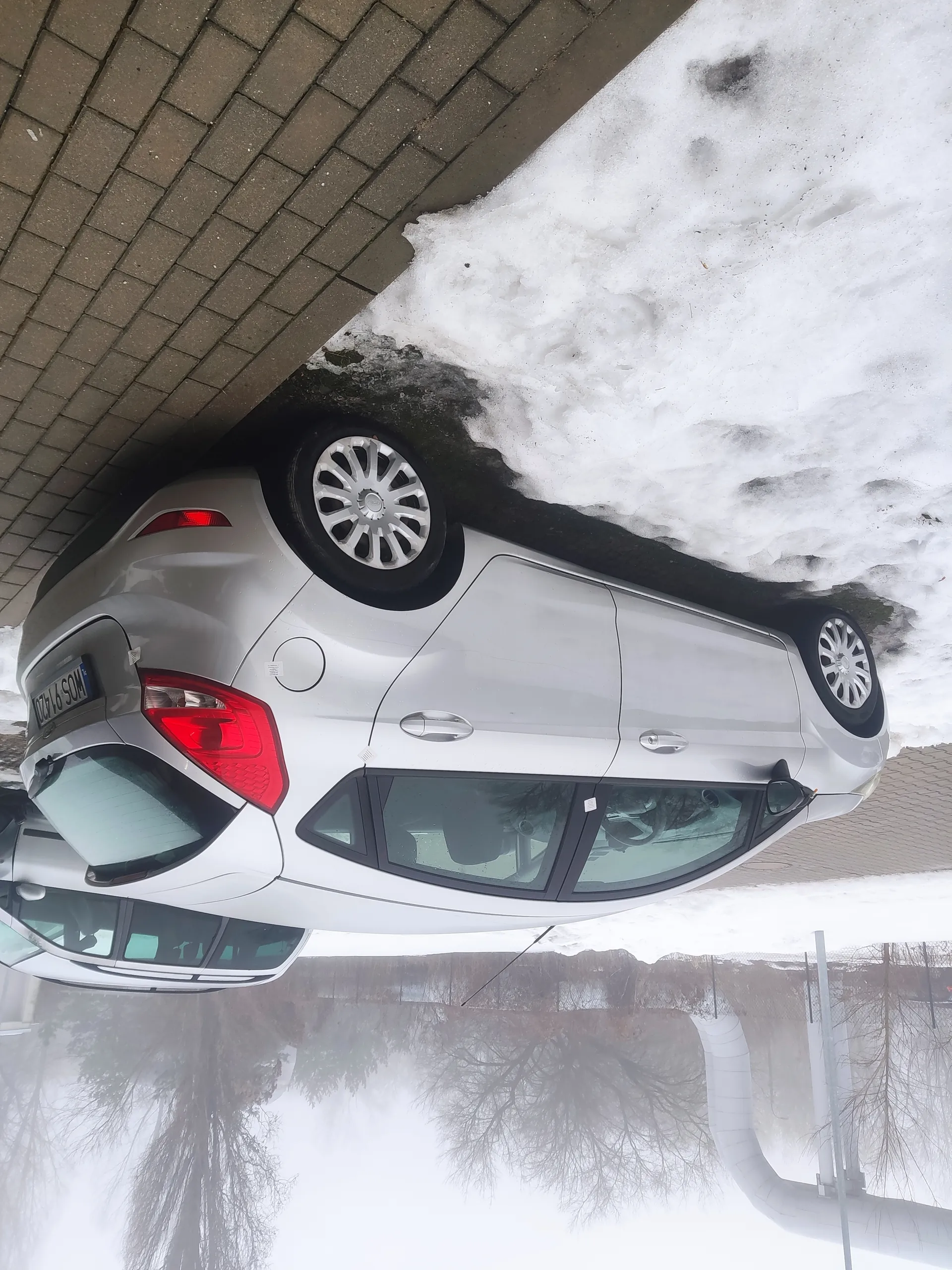 Srebrny samochód osobowy typu hatchback zaparkowany przy ceglanej ścianie, z widoczną tablicą rejestracyjną HOS 9420. Na ziemi śnieg, w tle mgła, drzewa bez liści i biały rurociąg. Widoczne czerwone światła tylnego oświetlenia oraz felgi aluminiowe.