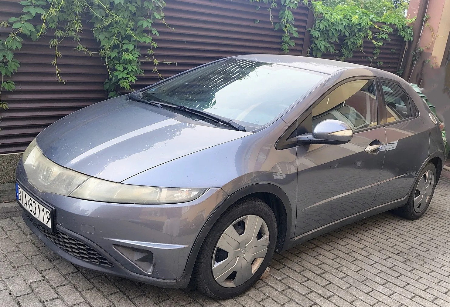 Srebrnoszary samochód osobowy marki Honda Civic w nadwoziu hatchback, zaparkowany na kostce brukowej. Auto posiada tablicę rejestracyjną o numerze BIA 83119. W tle znajduje się brązowy, poziomo żebrowany płot, nad którym zwisają zielone rośliny pnące.