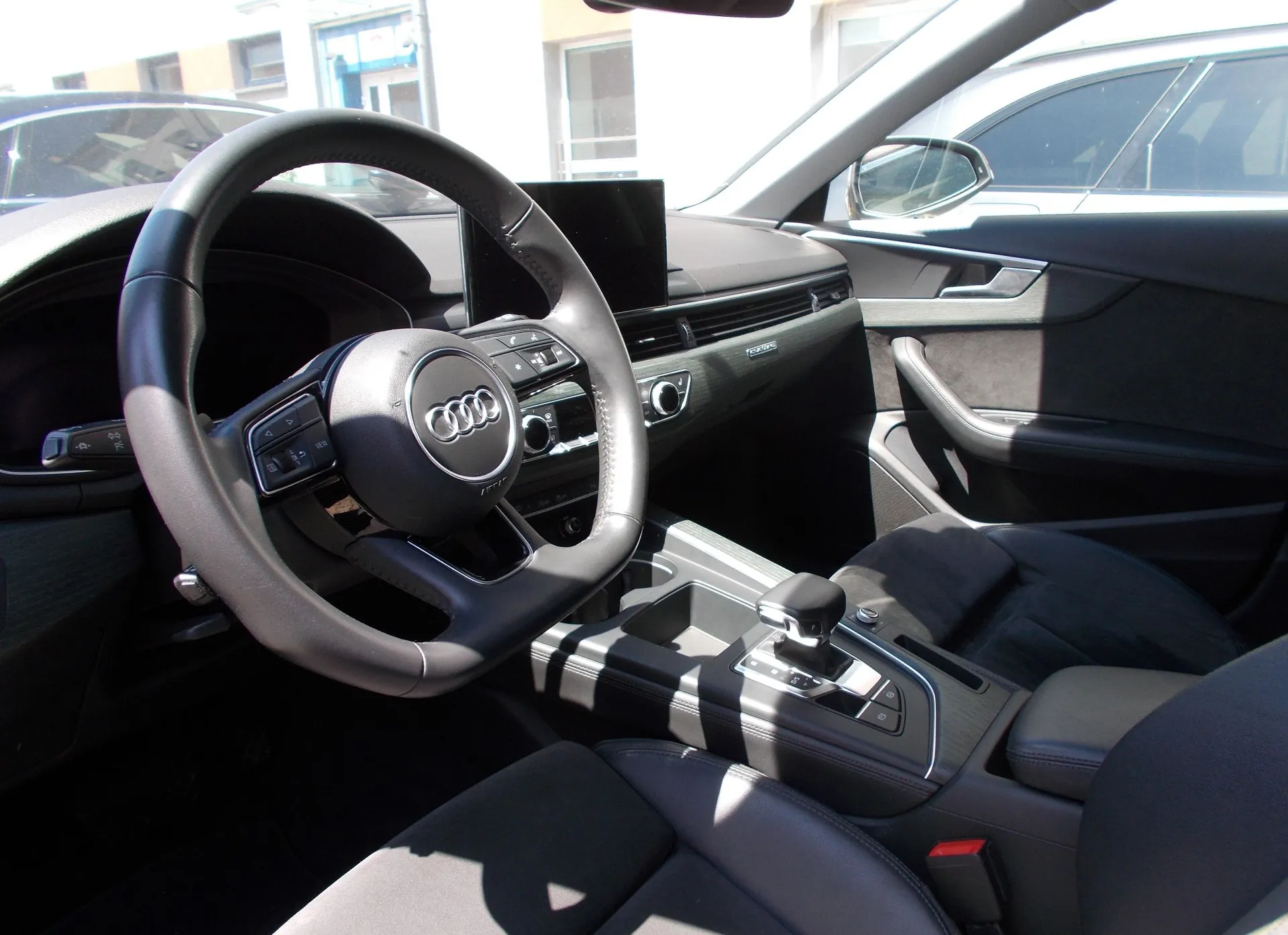 Wnętrze samochodu marki Audi z perspektywy kierowcy. Widoczna jest czarna, skórzana kierownica z logo Audi w centrum, deska rozdzielcza z ekranem multimedialnym, konsola środkowa z dźwignią zmiany biegów oraz ciemne fotele. W tle, przez przednią szybę i lusterko boczne, widoczne są fragmenty innych pojazdów i budynków.