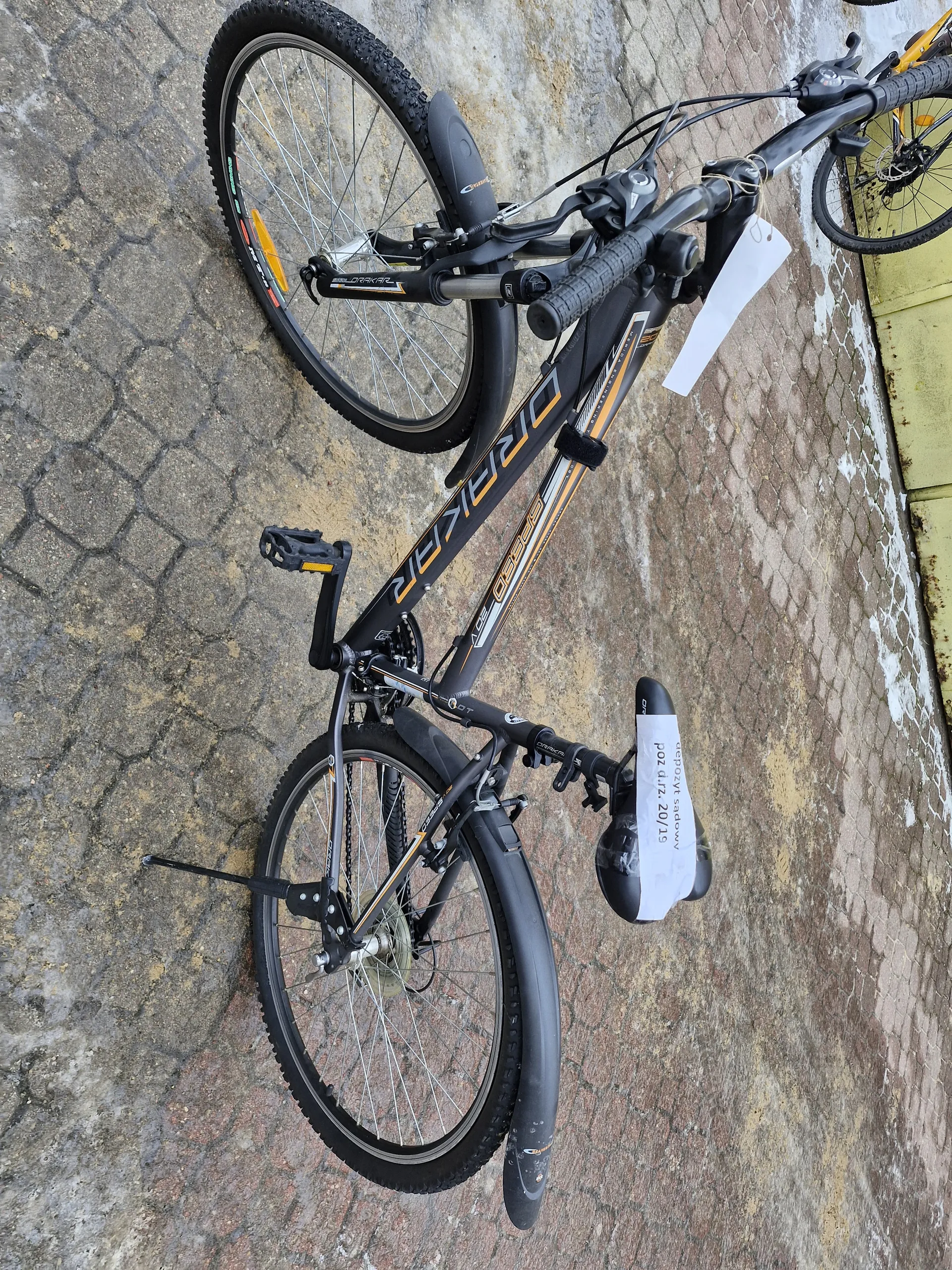 Czarny rower marki DRAKAR z pomarańczowymi akcentami, stojący na brukowanej powierzchni. Na siedzeniu widoczny biały napis: 'depozyt sądowy poz d.rz. 20/19'. W tle fragment żółtej ściany i części innego roweru. Rower posiada czarne opony, kierownicę z dźwigniami oraz bagażnik przedni.