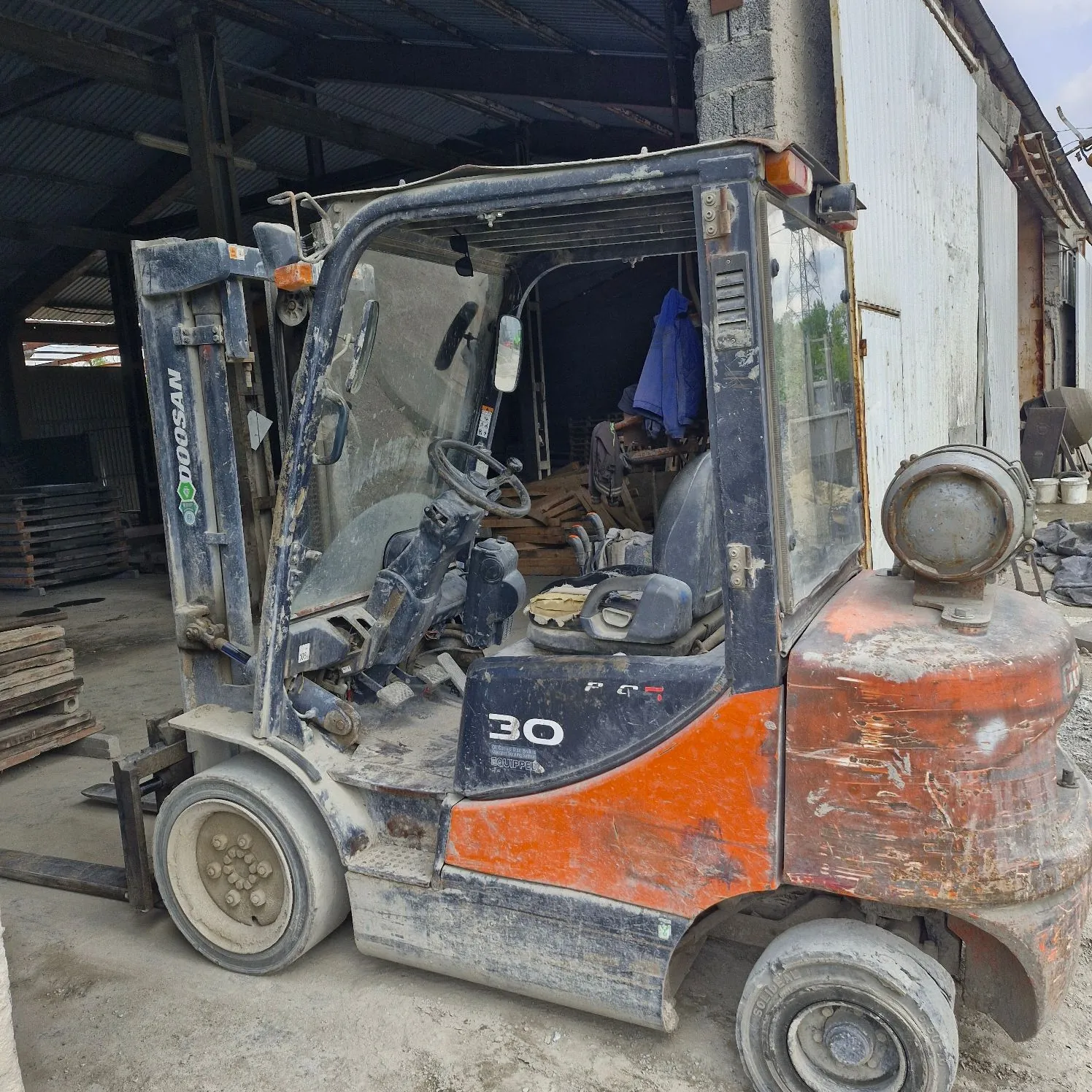 Stary, zakurzony wózek widłowy marki Doosan w kolorach pomarańczowym i szarym, zaparkowany wewnątrz otwartego budynku przemysłowego. Na boku maszyny widnieje napis 'DOOSAN' oraz liczba '30'. Pojazd jest w złym stanie technicznym, z widocznymi śladami rdzy i zabrudzeń.