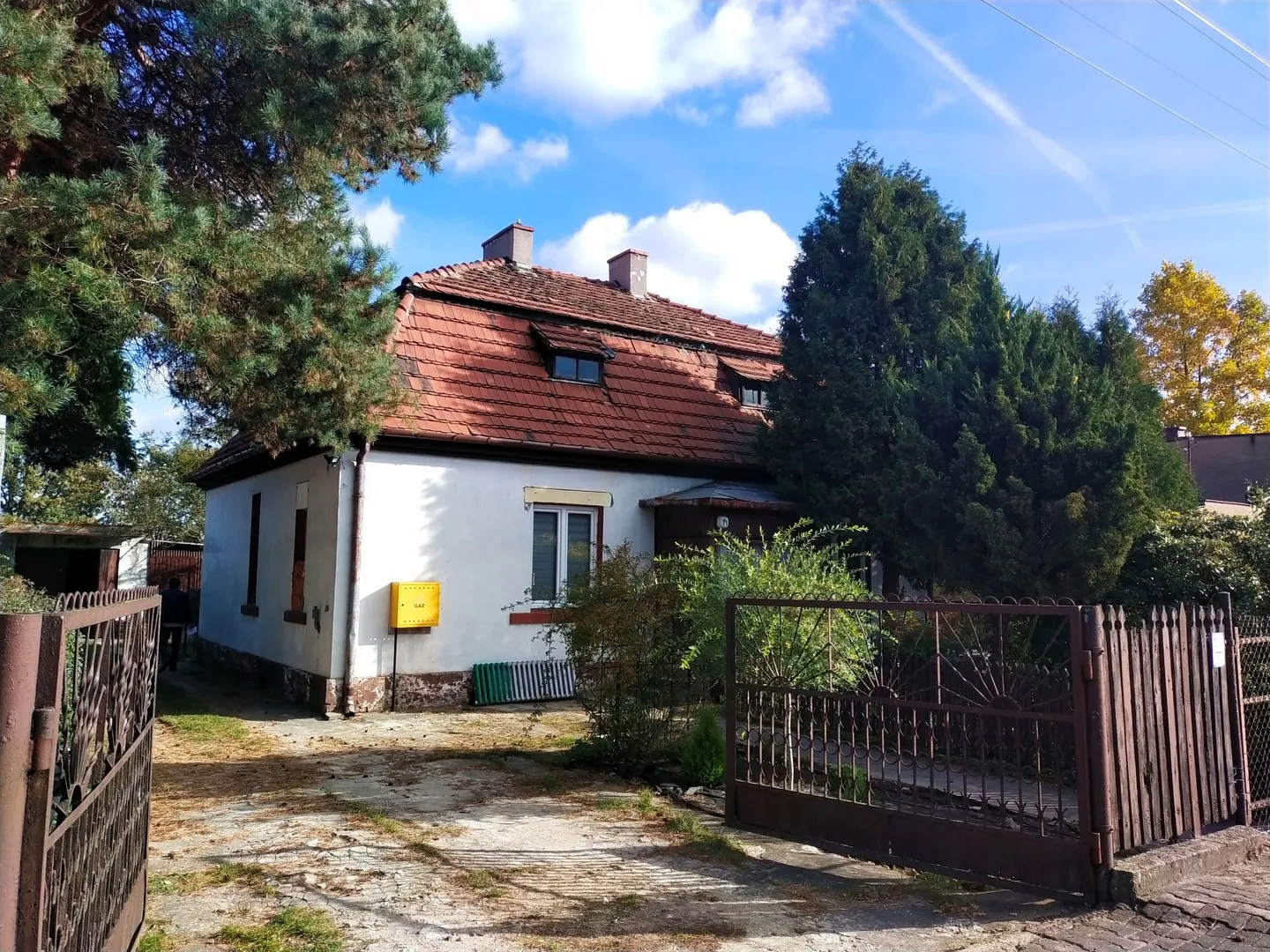 Jednopiętrowy biały dom z czerwonym, spadzistym dachem krytym dachówką. Na ścianie budynku znajduje się żółta skrzynka pocztowa. Dom otoczony jest zielenią, w tym dużymi, gęstymi drzewami iglastymi po prawej stronie i częścią drzewa po lewej. Przed domem znajduje się podjazd z betonowych płyt i ziemi, ograniczony brązową, metalową bramą po prawej stronie oraz fragmentem ogrodzenia po lewej. W tle widać niebieskie niebo z białymi chmurami.