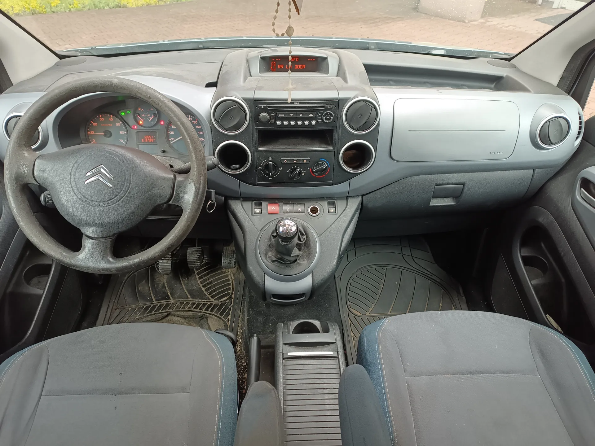 Wnętrze samochodu Citroën z widokiem na deskę rozdzielczą, kierownicę z logo marki, wskaźniki prędkości i obrotomierze, centralny panel z radioodtwarzaczem, przyciskami i gałkami sterującymi, dźwignią zmiany biegów oraz siedzeniami w kolorze szarym z niebieskimi akcentami. Na desce rozdzielczej widoczny wyświetlacz z czerwonymi cyframi, a na lusterku wstecznym wiszący krzyżyk. Podłoga pokryta brudnymi matach. Przez przednią szybę widoczna brukowana droga i zieleń.