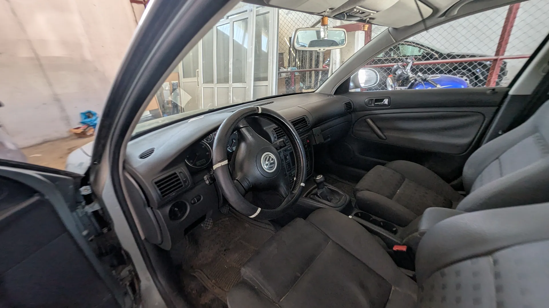 Wnętrze samochodu marki Volkswagen z otwartą drzwiami po stronie kierowcy. Widoczne czarne, zabrudzone siedzenia, kierownica z logo VW, deska rozdzielcza z licznikami i przyciskami, dźwignia zmiany biegów. W tle przez przednią szybę i okno widać garaż z siatką ochronną, białe drzwi, niebieski motocykl oraz fragmenty innych pojazdów. Podłoga w samochodzie pokryta brudnymi dywanikami.