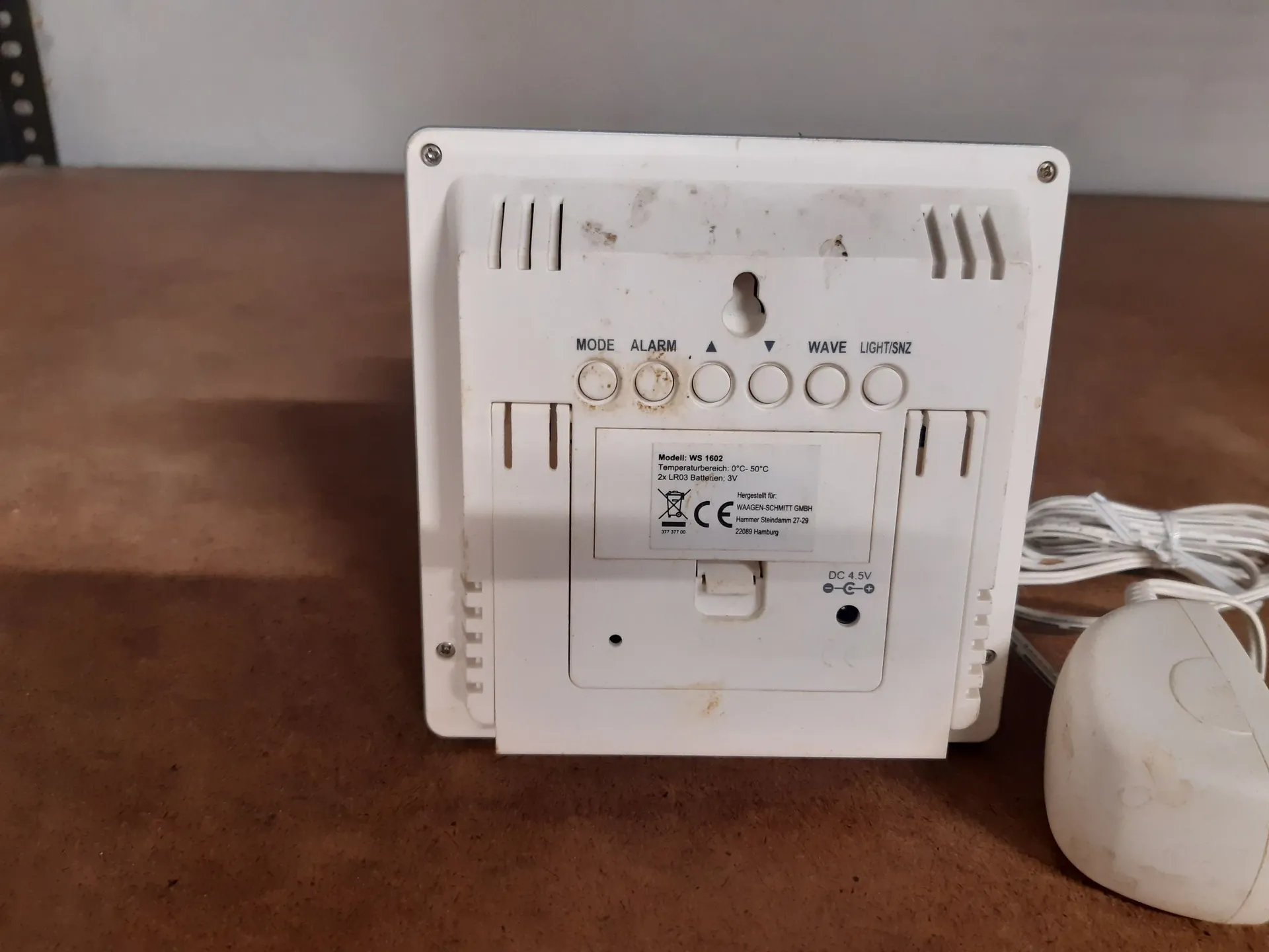 Tył białego, plastikowego urządzenia elektronicznego, prawdopodobnie zegara lub stacji pogodowej. Na obudowie znajdują się przyciski z napisami: MODE, ALARM, strzałka w górę, strzałka w dół, WAVE oraz LIGHT/SNZ. Poniżej znajduje się tabliczka znamionowa z tekstem: 'Modell: WS 1502, Temperaturbereich: 0°C-50°C, 2x LR03 Batterien, 3V', logo CE oraz informacja o producencie: 'Hergestellt für WAGEN SCHMITT GmbH, Hammer Straße 21-29, 22399 Hamburg'. W dolnej części widoczne jest gniazdo zasilania z opisem 'DC 4.5V'. Obok urządzenia leży biały kabel i okrągły czujnik.