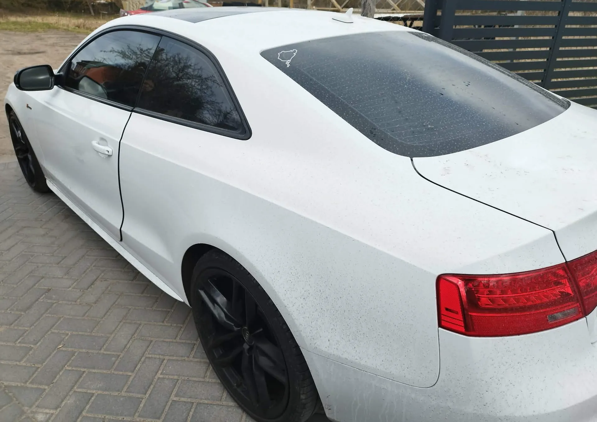 Biały samochód osobowy typu coupe zaparkowany na kostce brukowej. Widok z tyłu i z boku pojazdu. Samochód ma przyciemniane szyby, czarne felgi oraz czerwoną tylną lampę. Na tylnej szybie znajduje się mała naklejka z konturem mapy Polski.