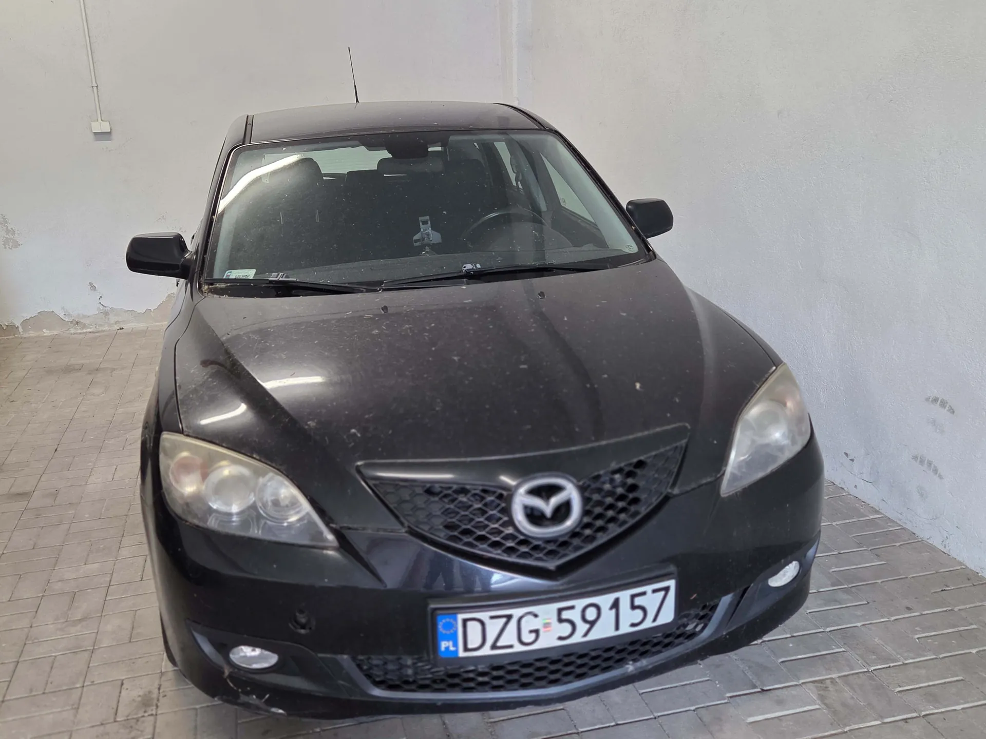 Czarny samochód marki Mazda zaparkowany w jasnym garażu. Widoczny jest przód pojazdu z logo Mazdy na grillu oraz tablicą rejestracyjną o numerze DZG 59157. Karoseria samochodu jest zakurzona.