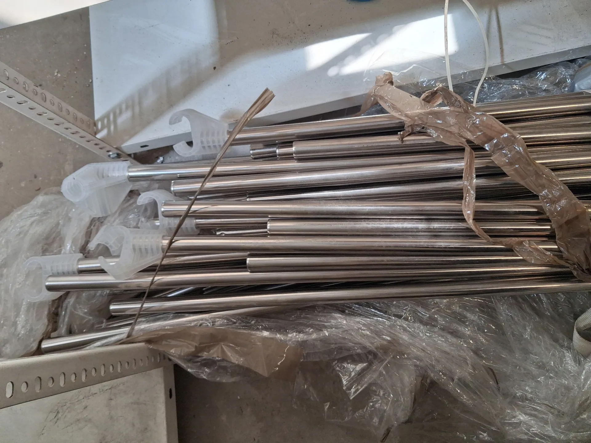 Zbiór metalowych, srebrnych rurek i prętów o różnych średnicach, ułożonych poziomo i częściowo przykrytych przezroczystą folią plastikową. Niektóre rurki mają na końcach białe, plastikowe nakładki. Całość znajduje się na metalowej półce z otworami montażowymi.