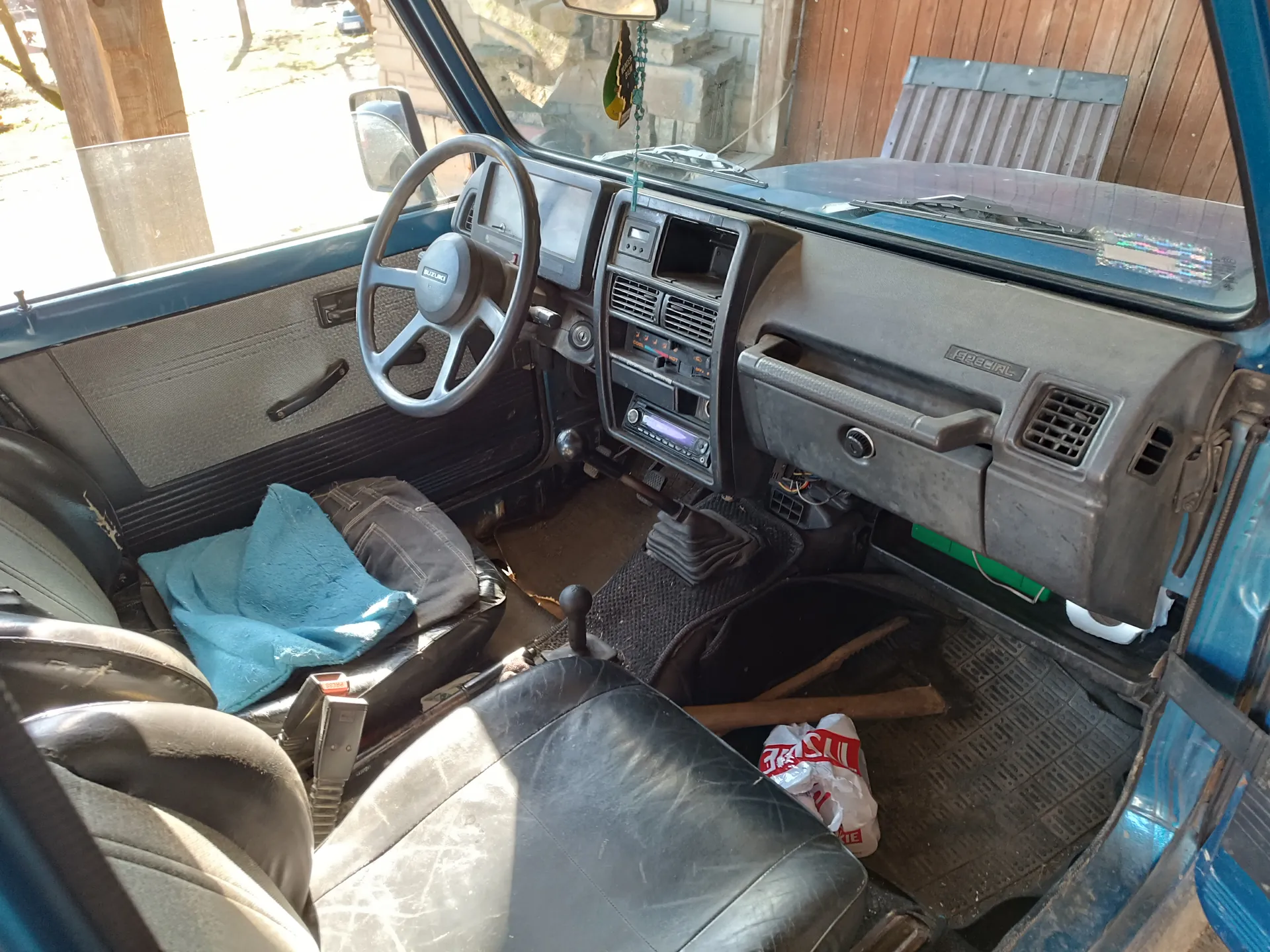 Wnętrze niebieskiego samochodu marki Suzuki. Widoczna kierownica z logo Suzuki, deska rozdzielcza z radioodtwarzaczem, przyciskami i wentylacjami. Na siedzeniu pasażera leży niebieska tkanina i ciemne spodnie. Na podłodze znajduje się plastikowa torba z czerwonym napisem 'LIE' oraz drewniany przedmiot. Czarne, zużyte siedzenia. Poza samochodem widoczny drewniany budynek i fragment podwórza. Na desce rozdzielczej widnieje napis 'SPECIAL'.