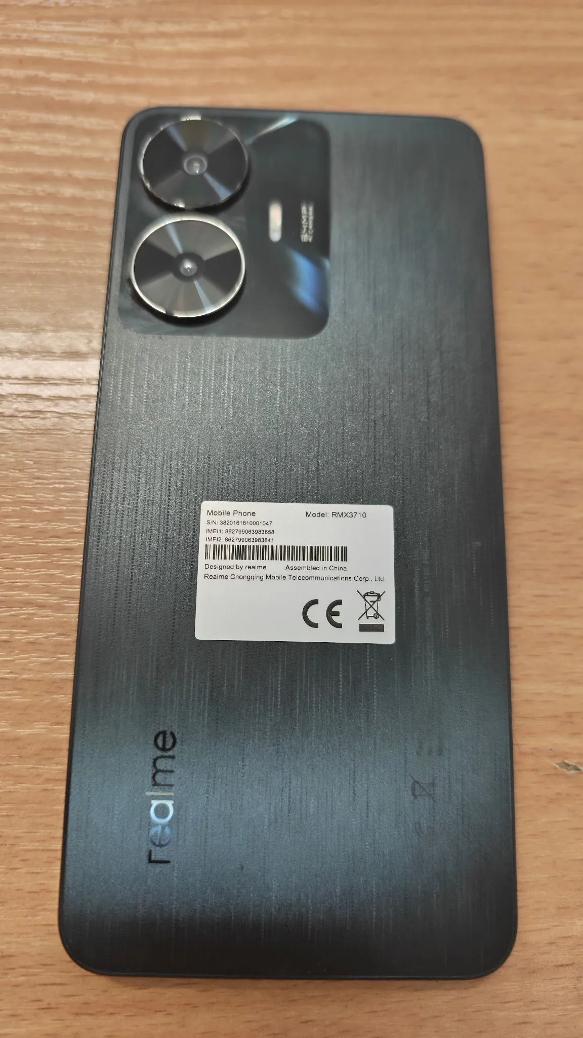 Tylna obudowa ciemnoszarego smartfona marki realme. W górnej części znajdują się dwa duże, okrągłe moduły aparatów. Na środku naklejona jest biała etykieta z danymi technicznymi: 'Mobile Phone', 'Model: RMX3710', numery seryjne S/N oraz IMEI, informacja 'Designed by realme', 'Assembled in China' oraz znak CE. W dolnej części obudowy widnieje pionowy napis 'realme'. Telefon leży na drewnianej powierzchni.