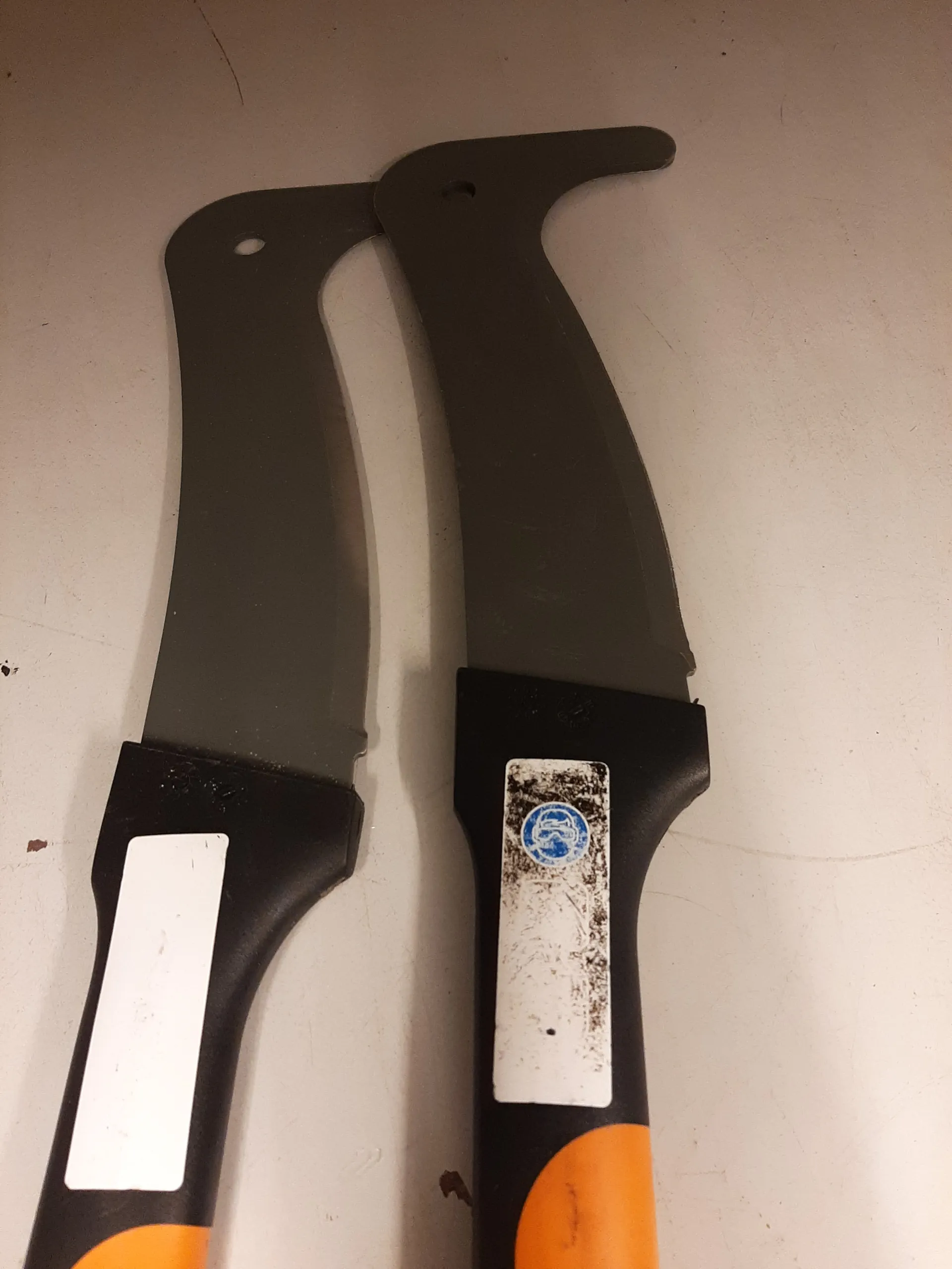 Dwa narzędzia ogrodnicze z czarnymi ostrzami i czarno-pomarańczowymi uchwytami. Każdy uchwyt posiada białą etykietę; na etykiecie po prawej stronie widoczny jest niebieski okrągły znak z białym symbolem. Narzędzia leżą na jasnej powierzchni.