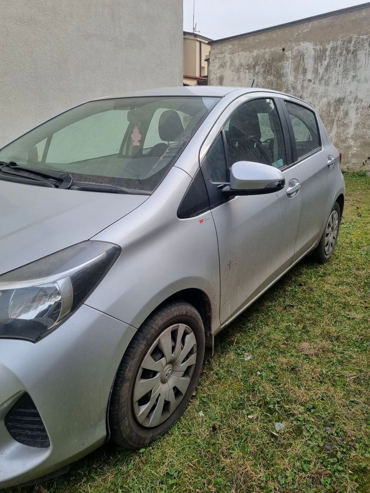 Srebrny samochód osobowy zaparkowany na trawie obok betonowej ściany budynku. Widoczne są przednie koło, lusterko boczne oraz czerwona naklejka na boku pojazdu. Wewnątrz samochodu, za przednią szybą, znajduje się mała naklejka w kształcie flagi Stanów Zjednoczonych. Trawa jest zielona z suchymi plamami. Ściana budynku ma zniszczoną, zatartą powierzchnię.