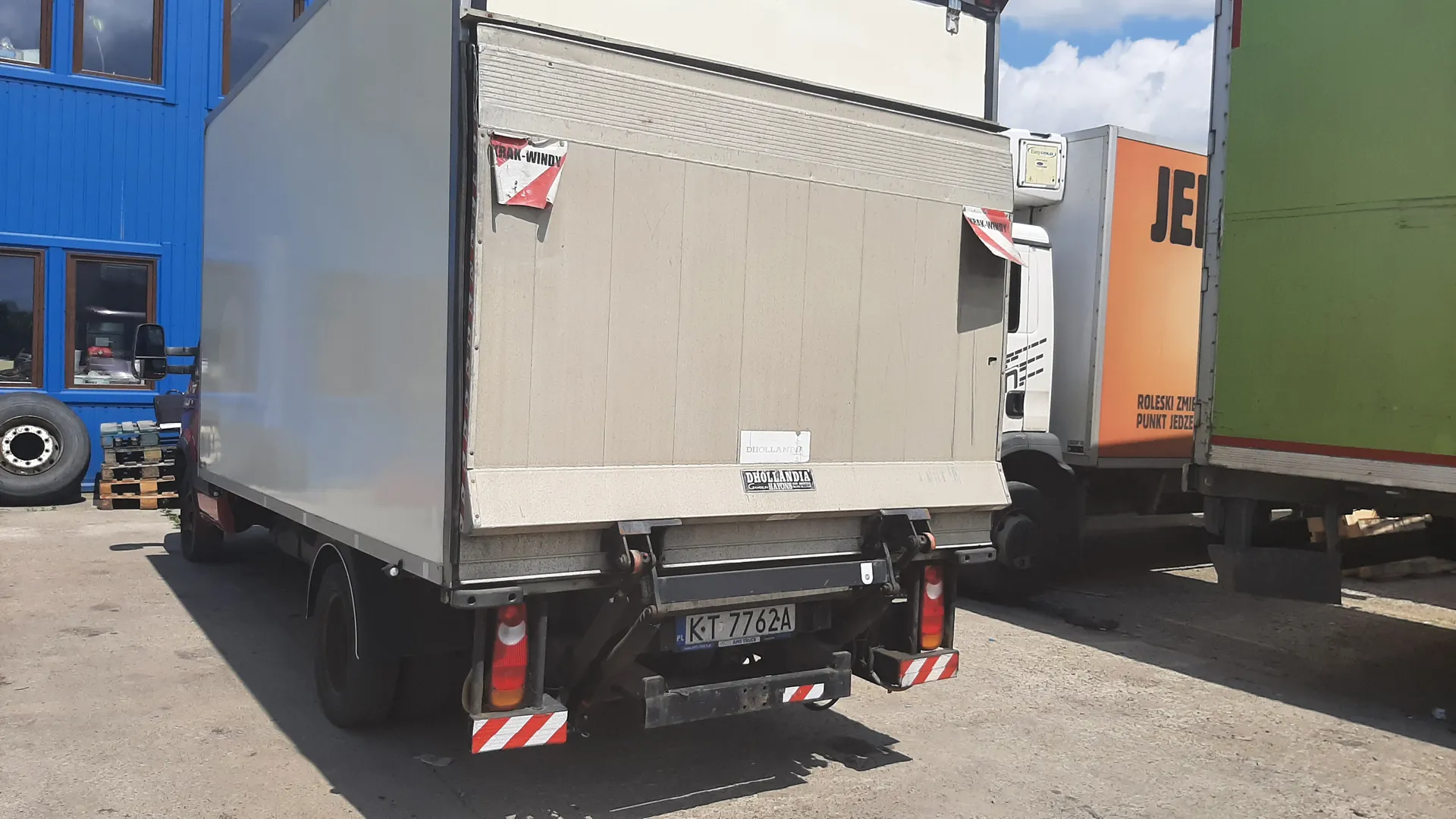 Tył beżowej ciężarówki z białym dachem, tablicą rejestracyjną KT 7762A, naklejkami 'TRAK-WINDY' (czerwono-białe) oraz 'DHOLLANDIA'. Widoczne czerwone światła i czerwono-białe odblaski. W tle niebieski budynek z oknami, opona, palety oraz inne ciężarówki: pomarańczowa z napisem 'JEL' i częściowo widocznym tekstem 'ROLESKI ZMIEN PUNKT JEDZIE' oraz zielona. Asfaltowa powierzchnia, niebo z chmurami.